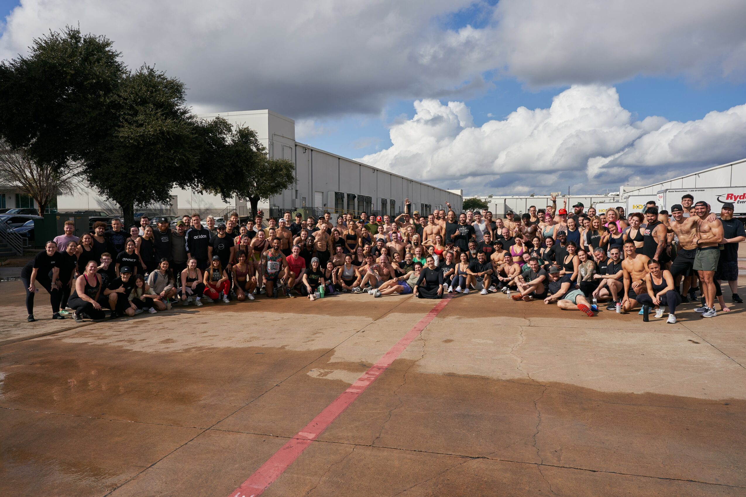 Large group of people gathered outdoors on a concrete surface with a building and cloudy sky in the background.