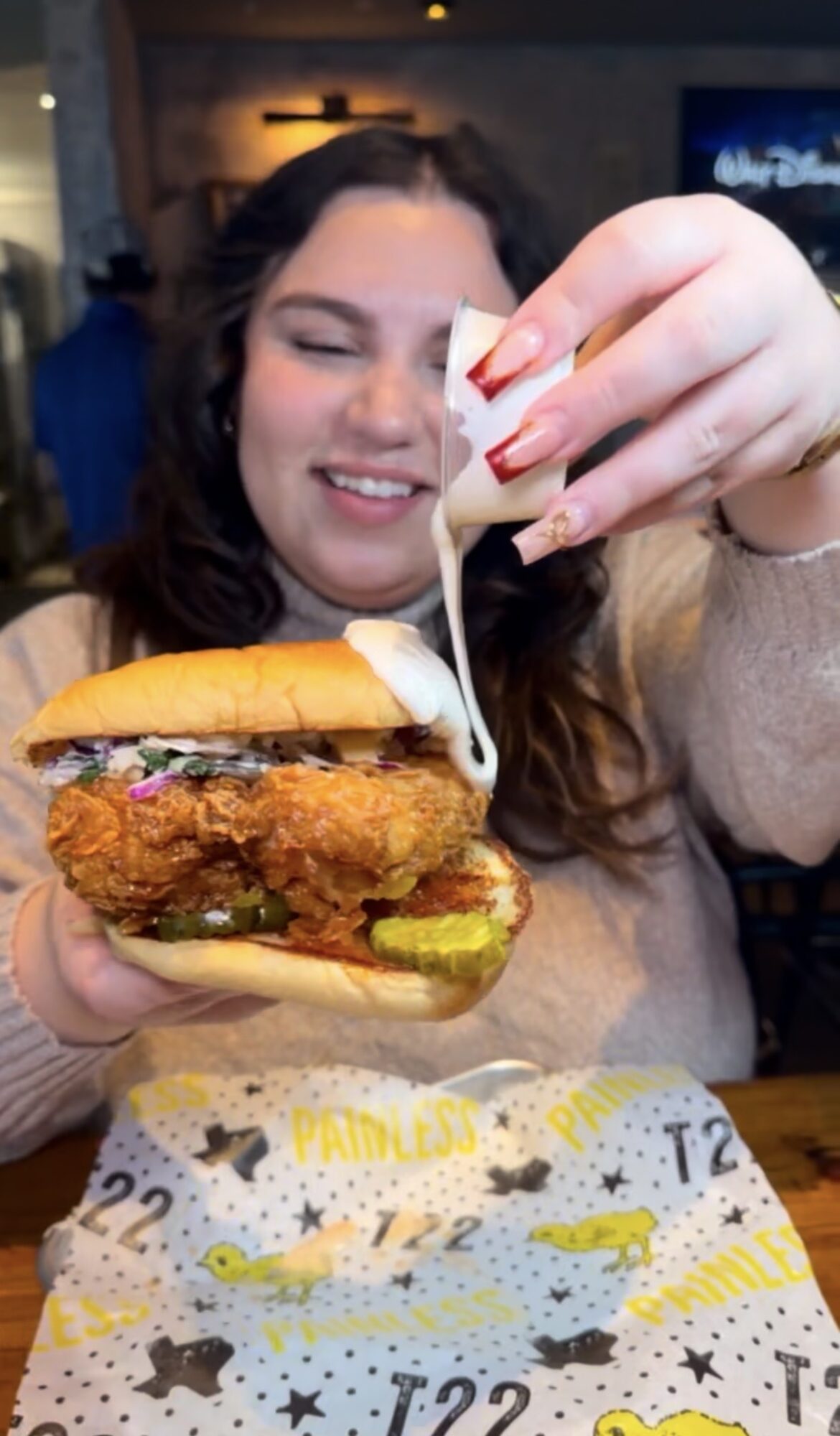 Person holding fried chicken sandwich with cheese, pickles, and sauce, smiling at a restaurant table.