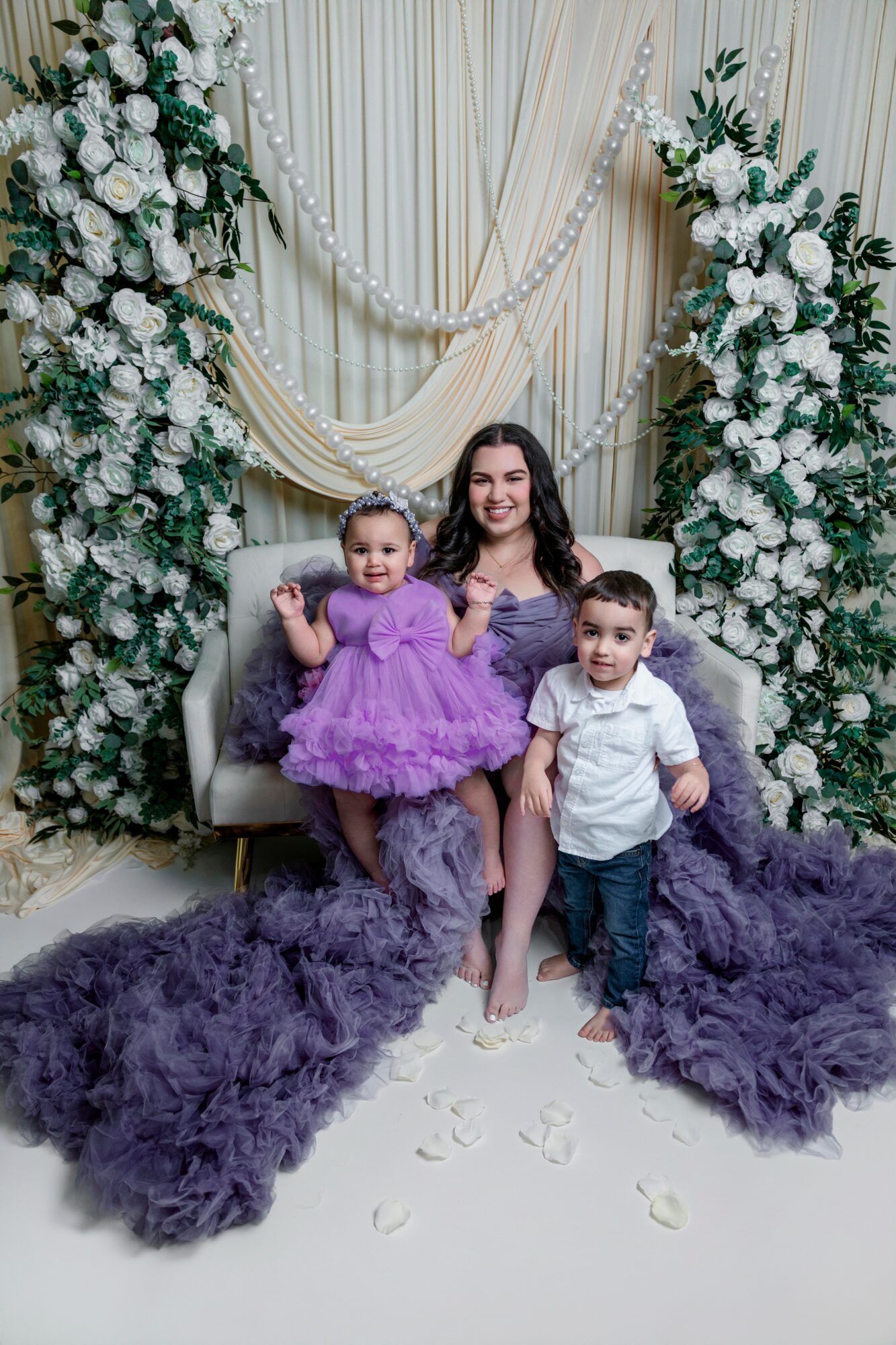 Woman with two children sitting on a decorated backdrop with white flowers and drapes, purple fabric on the floor, and flower petals.