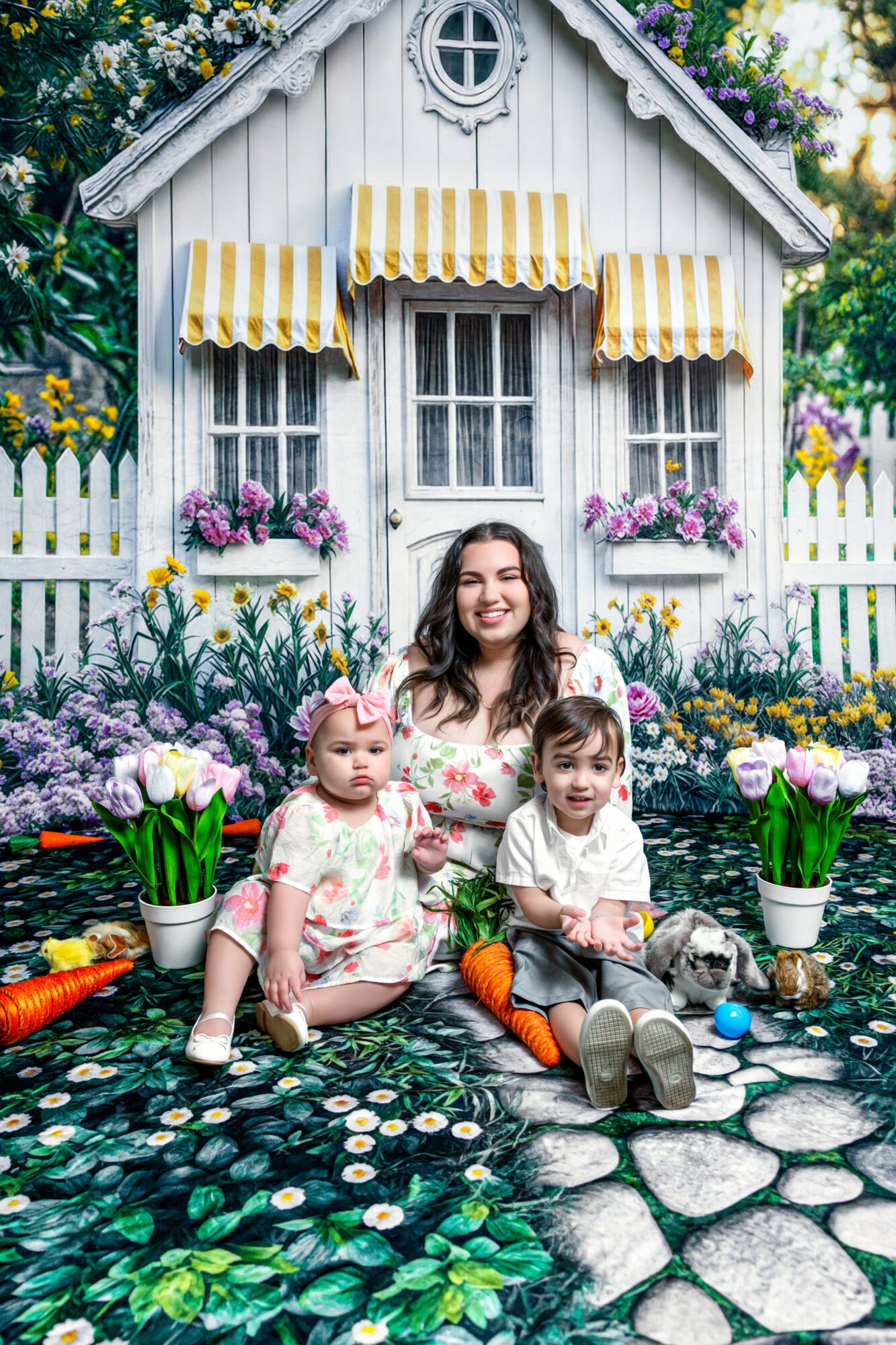 Woman with two children sitting on a stone path in front of a white house with yellow-striped awnings, surrounded by flowers.