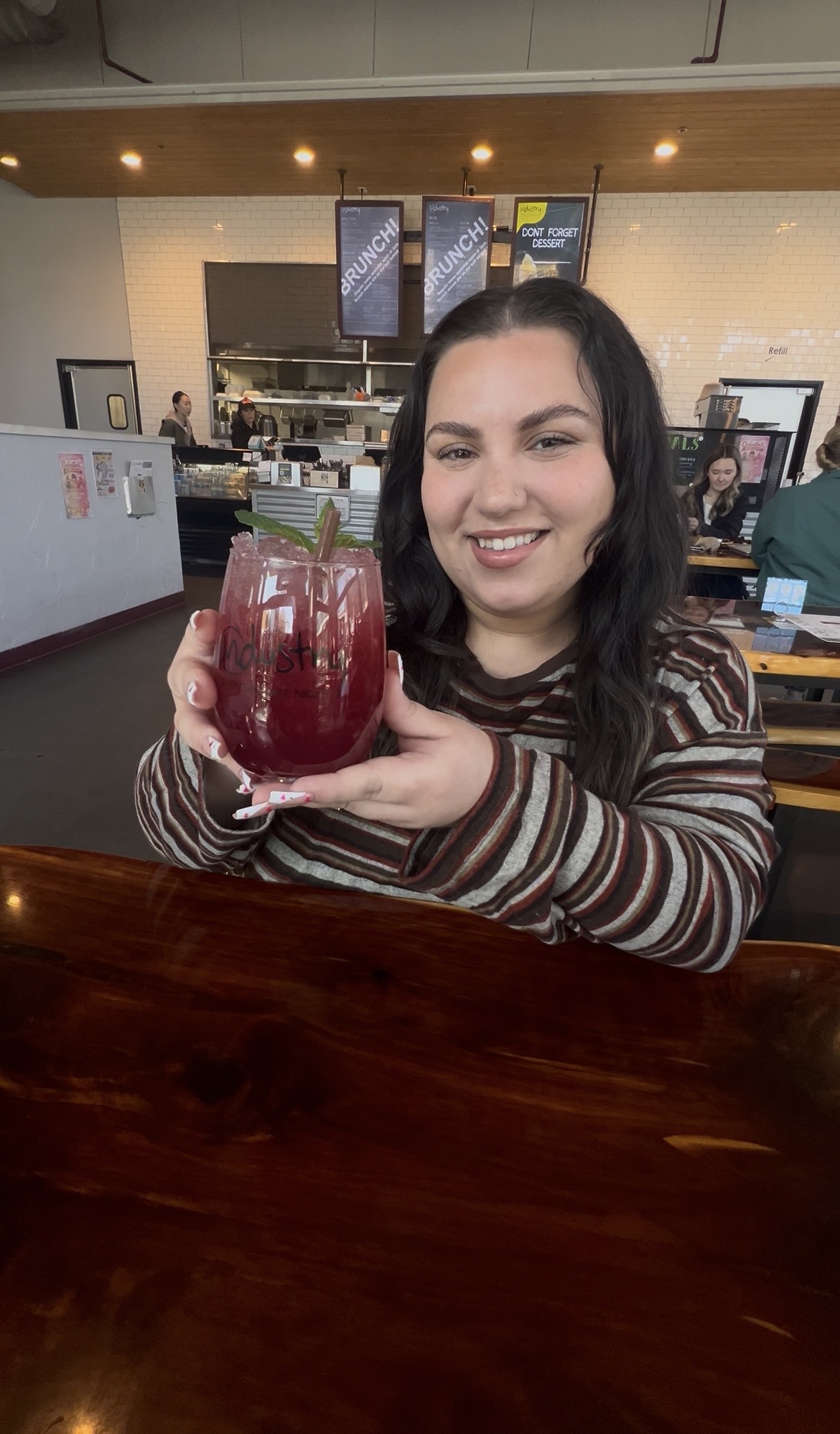 Smiling woman holding a large glass of red beverage in a cafe, with menu screens and counter in background.
