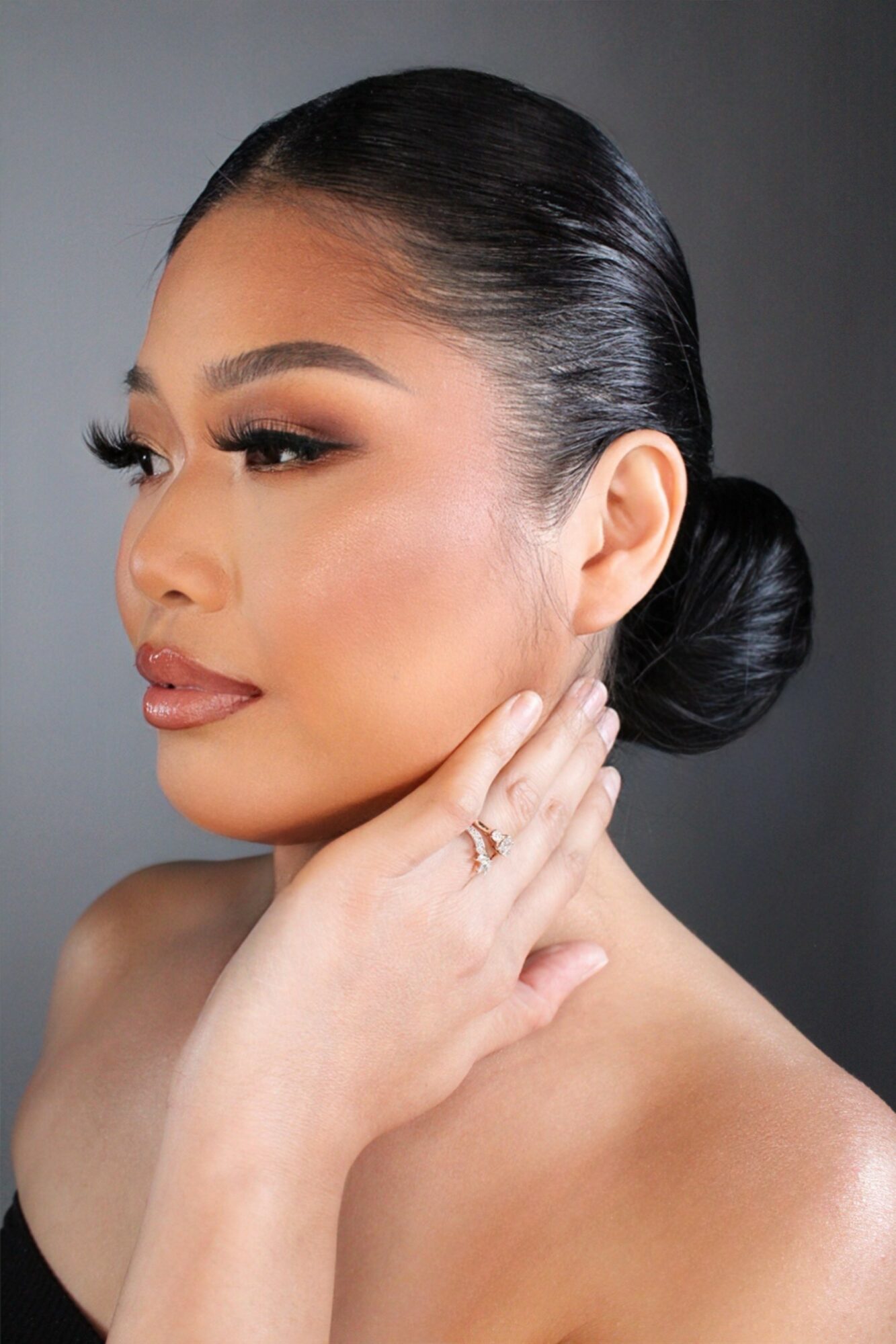 Close-up of a woman with sleek black hair in a bun, makeup, and a hand near her face, against a gray background.