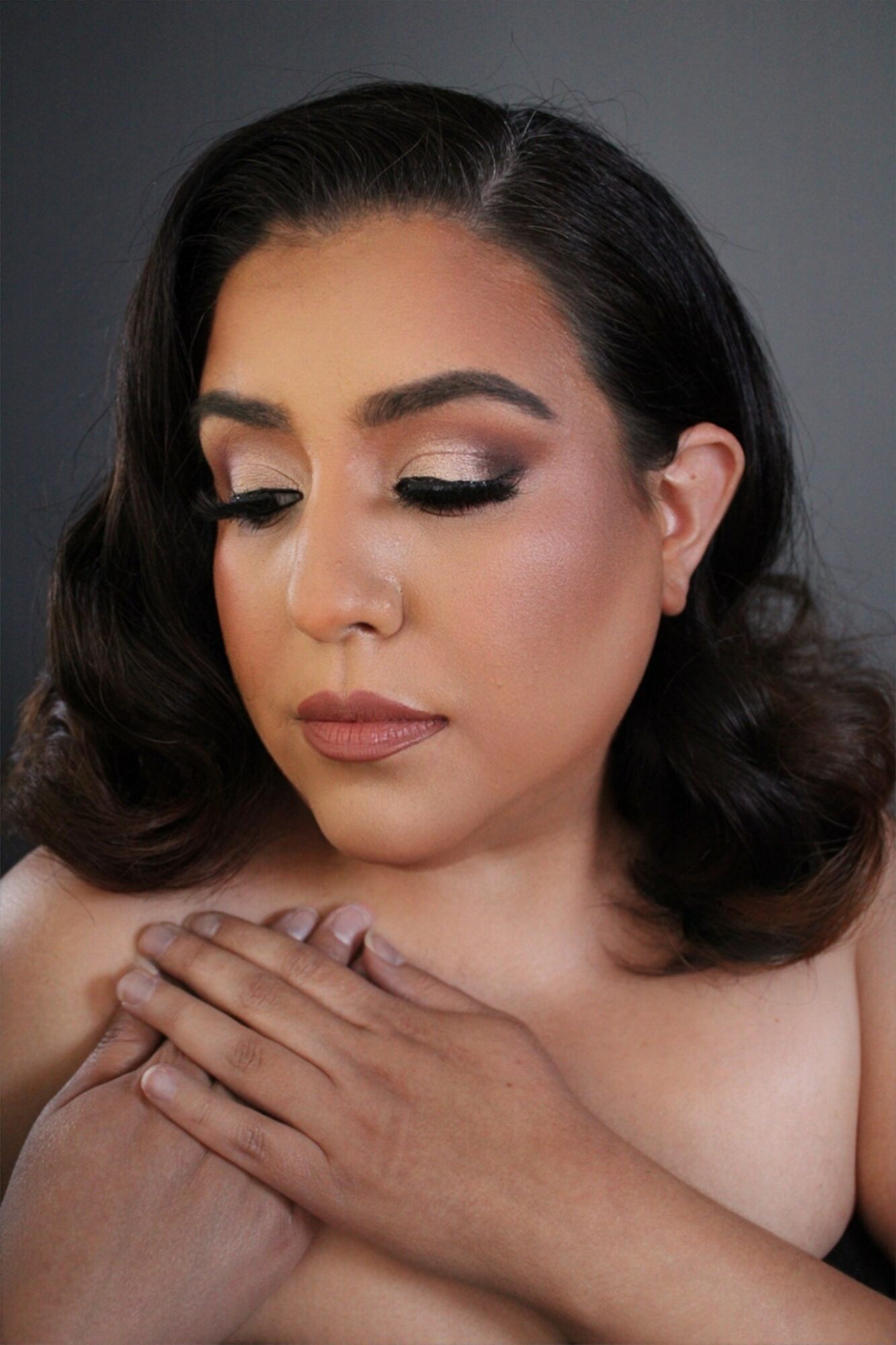 Close-up of a woman with makeup, eyes closed, hands crossed over chest, dark hair, neutral background.
