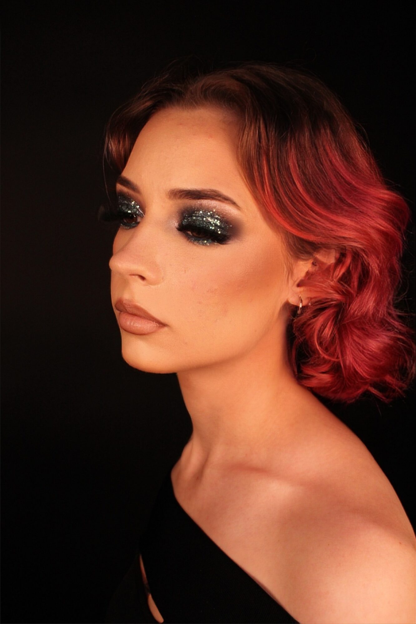 Woman with red and black wavy hair, makeup, and dark background, facing slightly to the left.