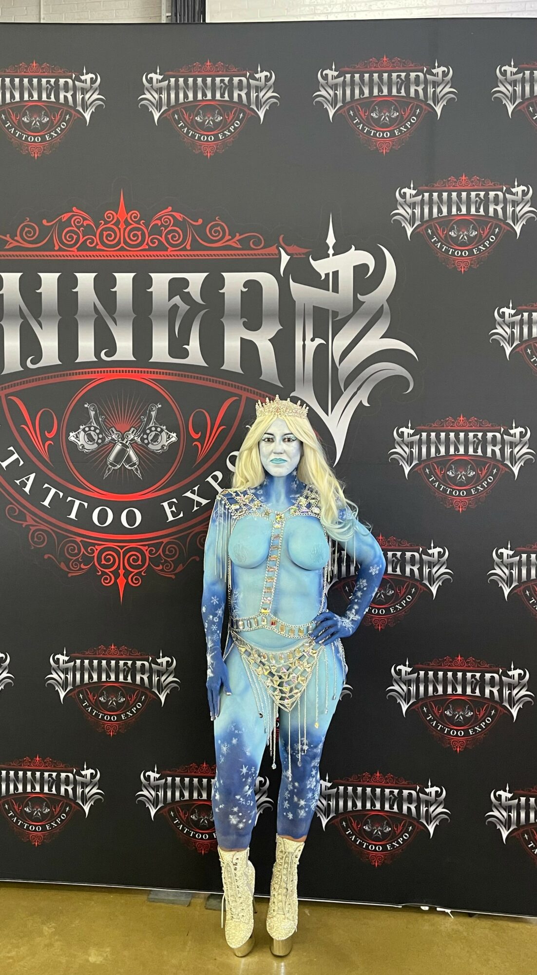 Woman with blue body paint, blonde hair, wearing beige boots, standing in front of a tattoo expo backdrop.