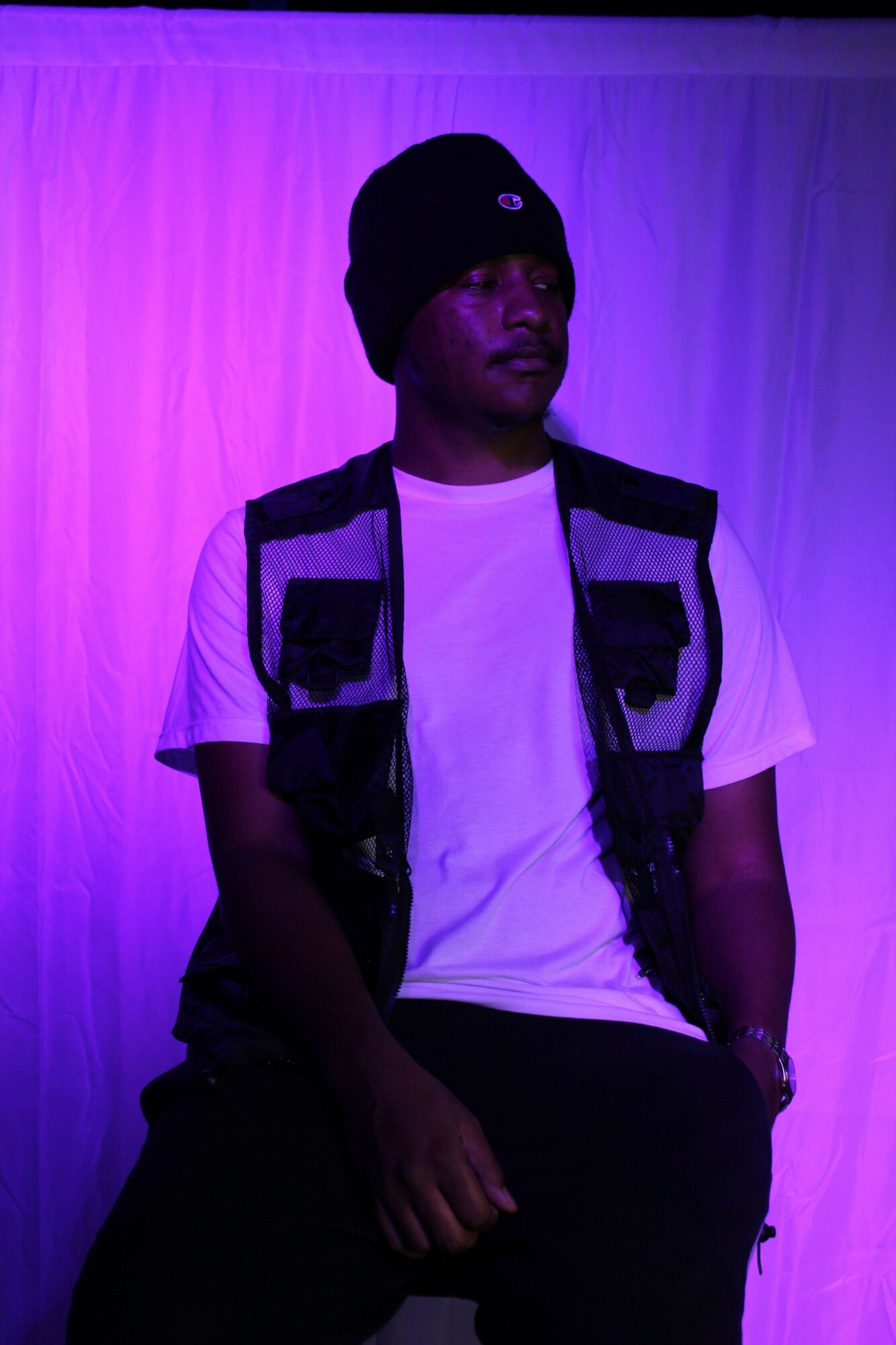 Person wearing a black cap, white shirt, and black jacket, sitting against a purple background.