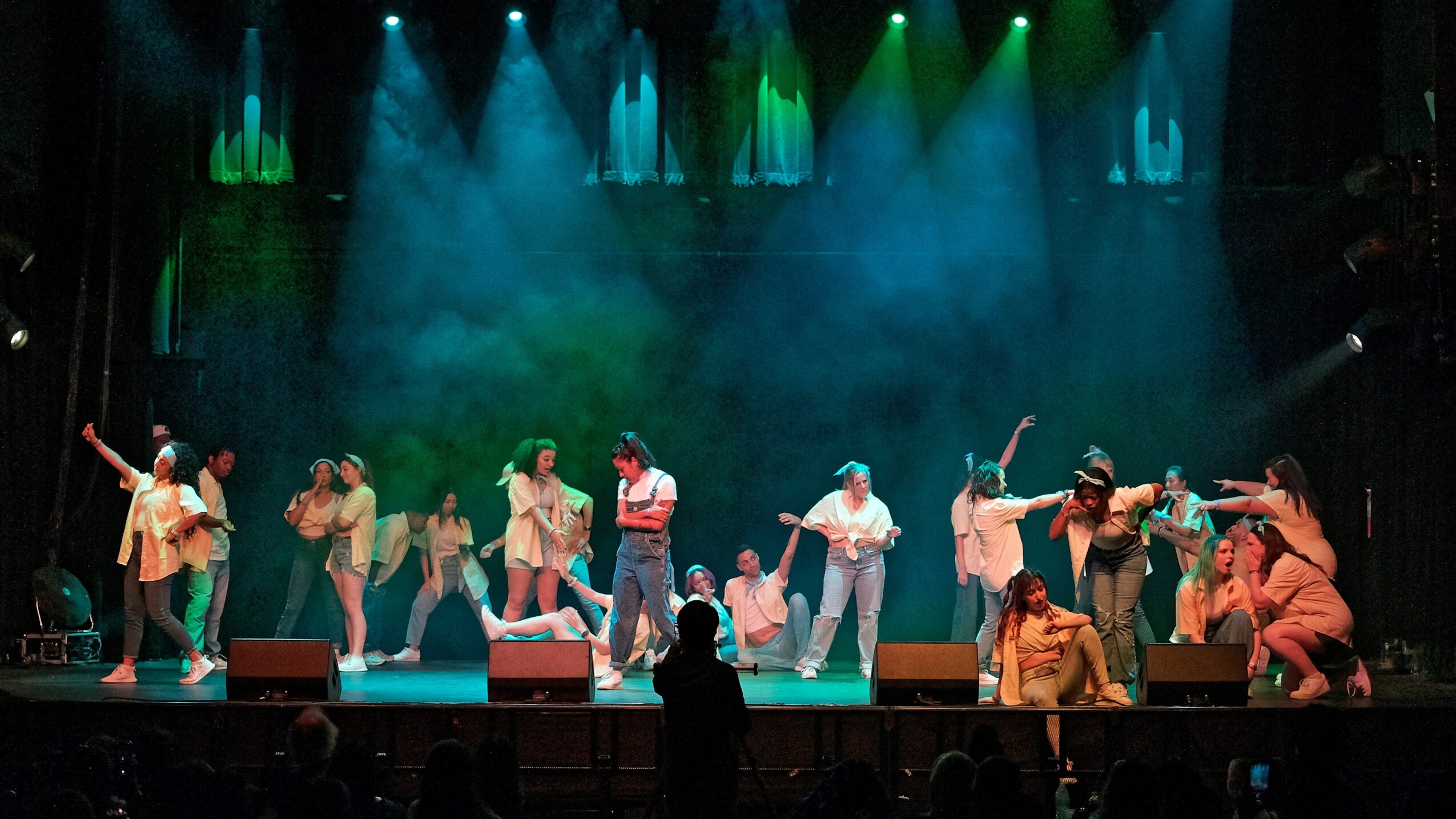 Group of people performing on stage with colorful lighting and smoke effects, some dancing and others standing.