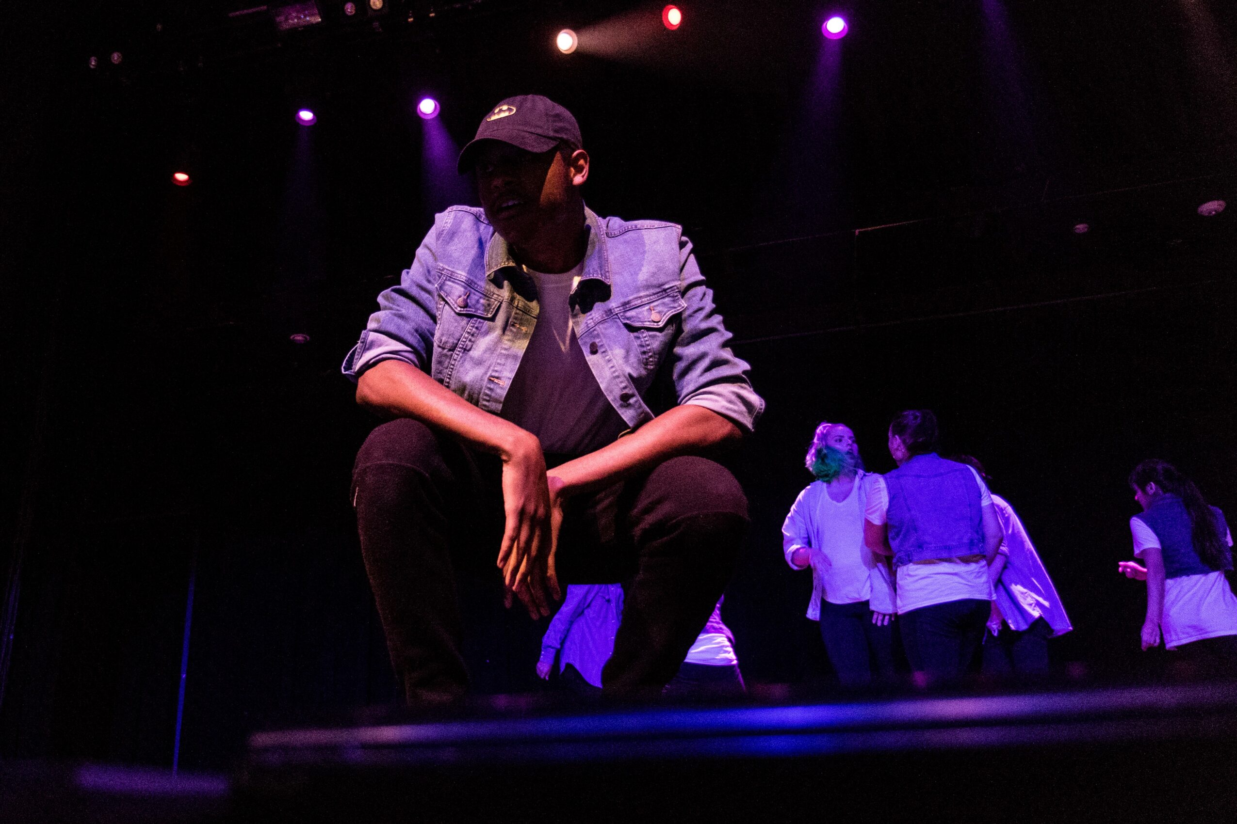 Person crouching on stage with purple and pink lights, wearing a denim jacket and cap, with others in the background.