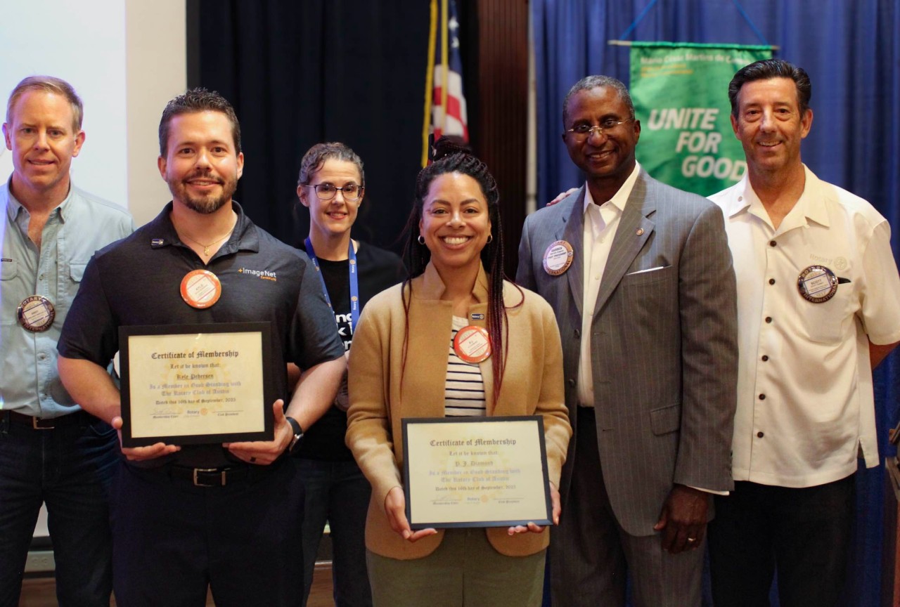Group of six people standing together, holding certificates, in front of a blue curtain and a green banner, smiling.