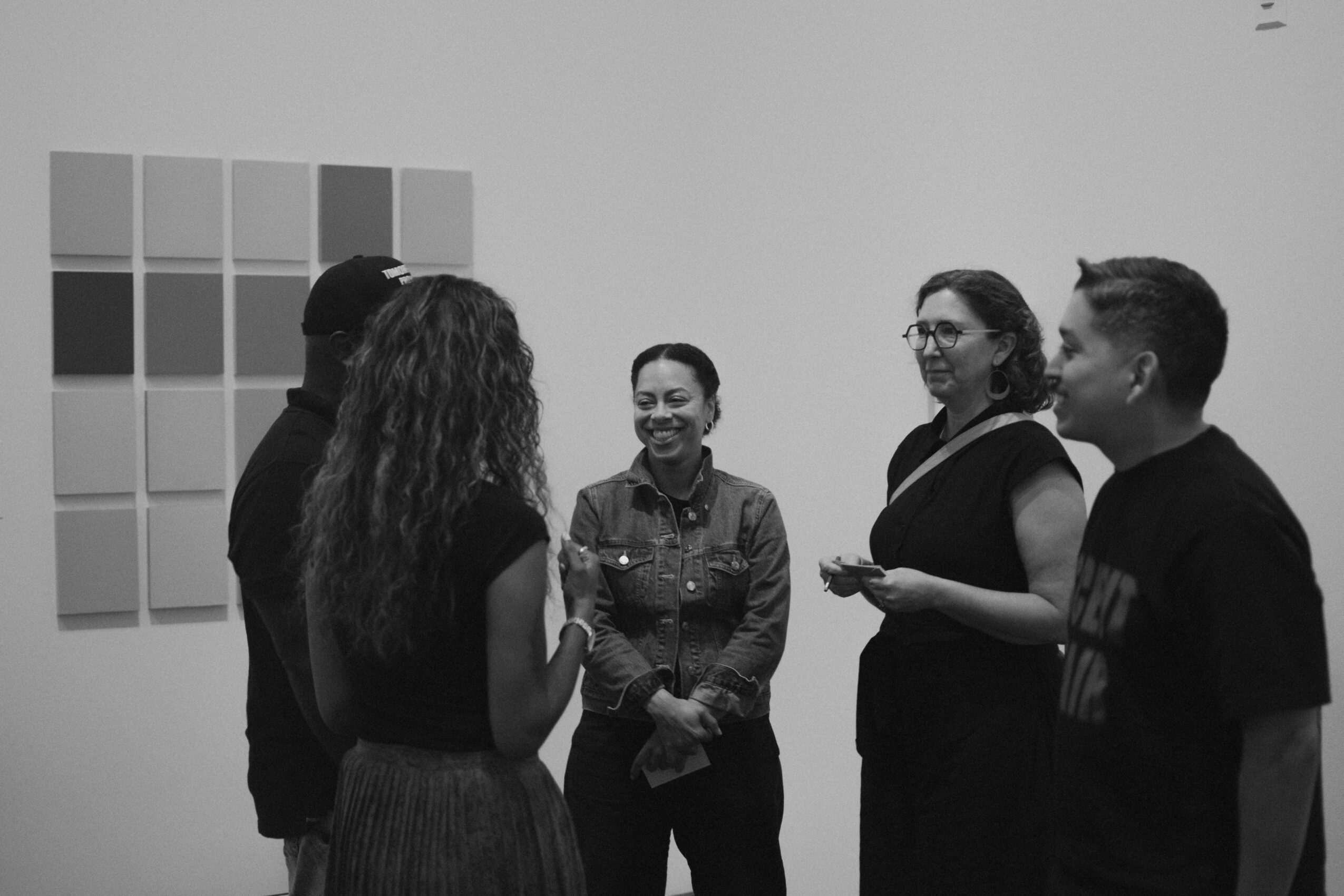 Four people standing and talking in an art gallery with a grid of colored squares on the wall.