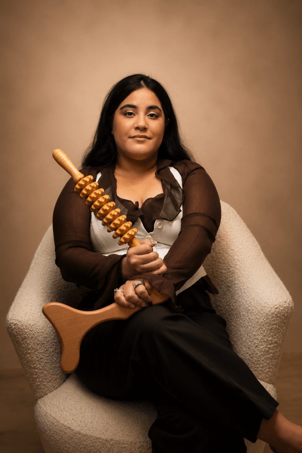Woman sitting in a beige chair holding a wooden object, wearing a black dress with sheer sleeves, against a plain brown background.