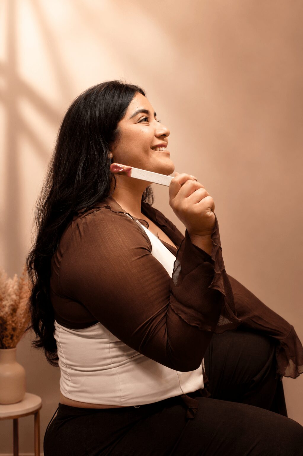 Woman with long dark hair smiling, holding a pink object near her face, sitting indoors with warm lighting.