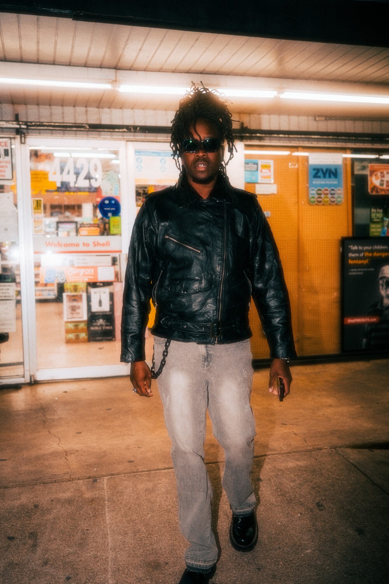 Person with curly hair and sunglasses wearing a black leather jacket and light pants standing outside a store at night.