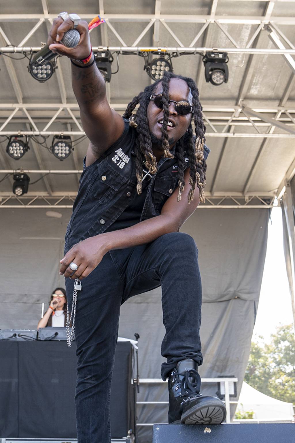 Performer with dreadlocks and sunglasses on stage, raising a hand, wearing a sleeveless vest and black pants, with stage equipment visible.