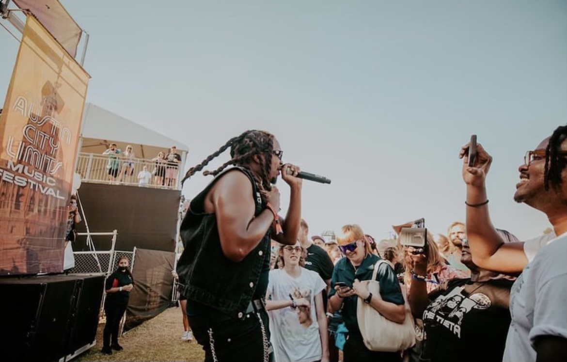 A woman with braided hair holds a microphone and performs outdoors with a crowd watching.