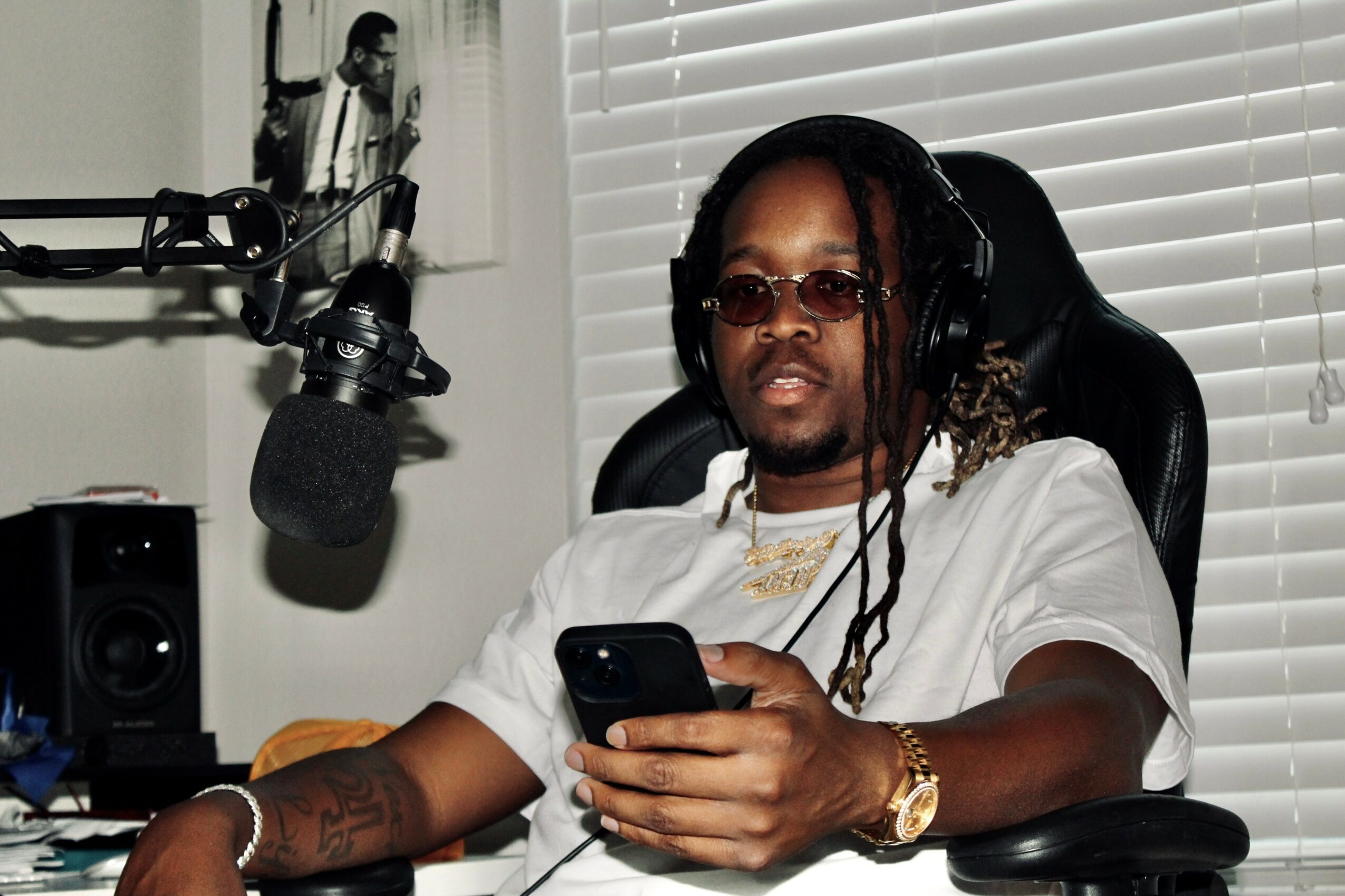 Person with sunglasses and dreadlocks sitting in a chair, holding a phone, in a room with blinds and audio equipment.