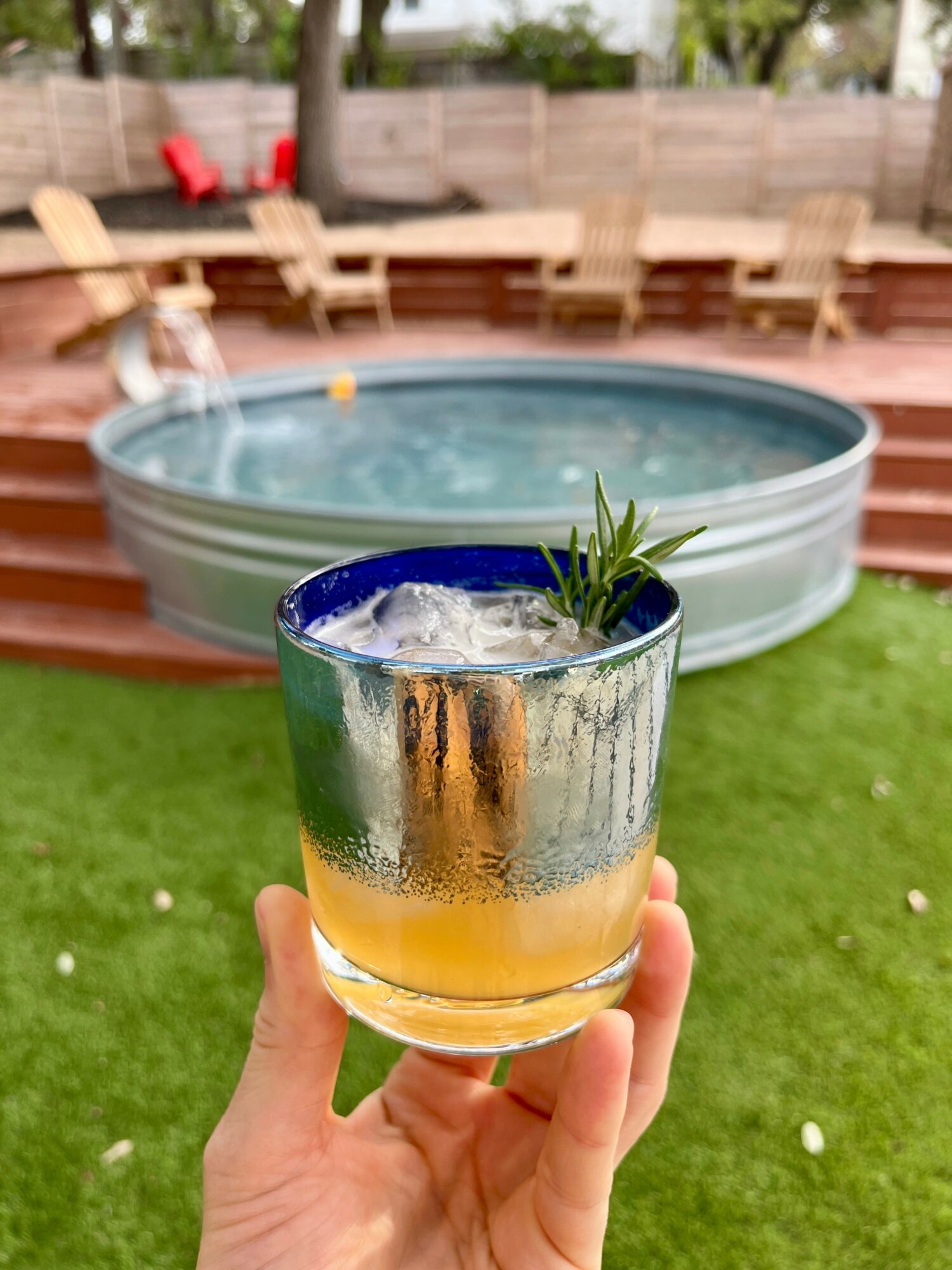 Hand holding a glass with a drink and a small plant, outdoor backyard with a hot tub and wooden deck, green grass.