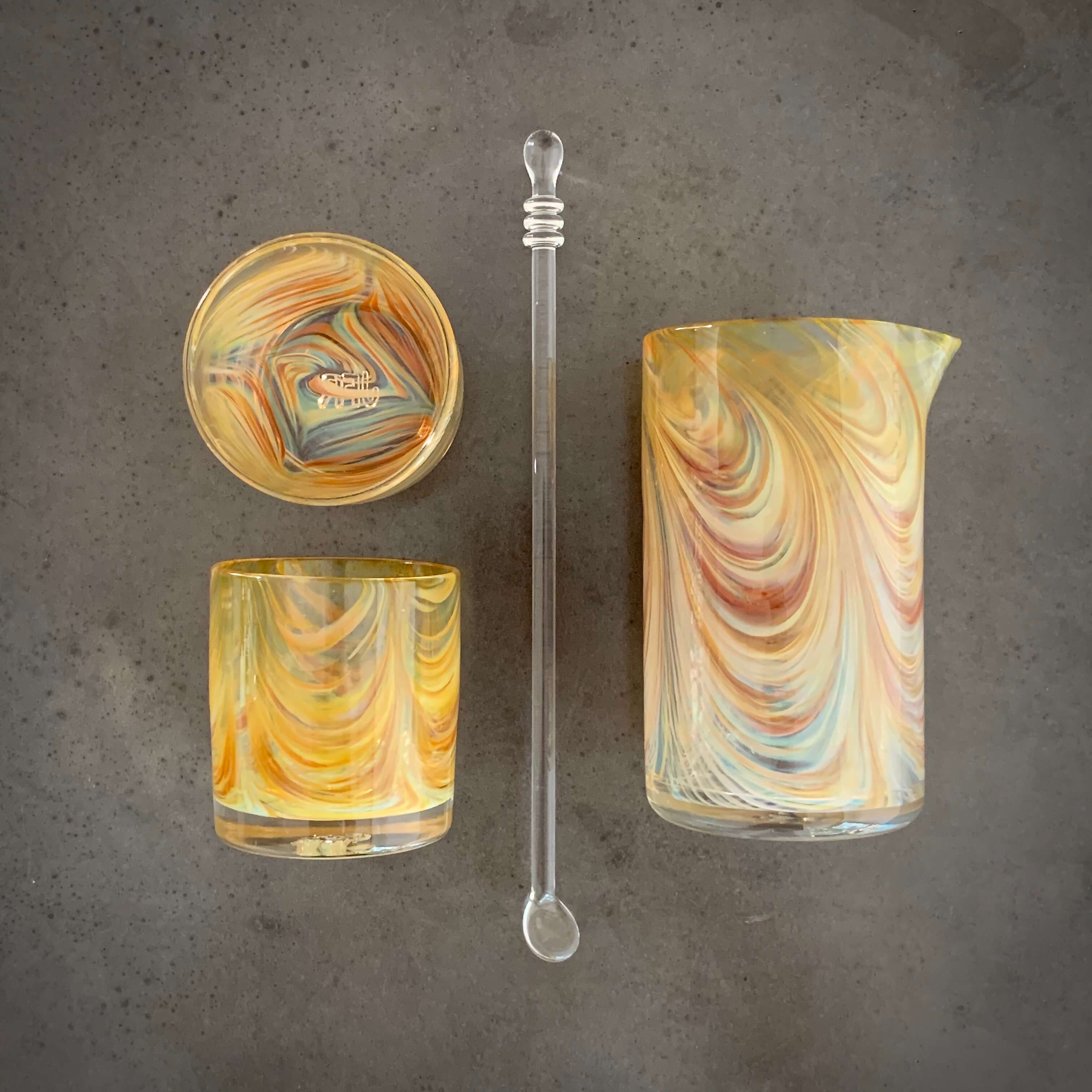 Three marbled glass objects and a glass dropper on a gray surface, arranged vertically. The objects include a bowl, a tumbler, and a pitcher.