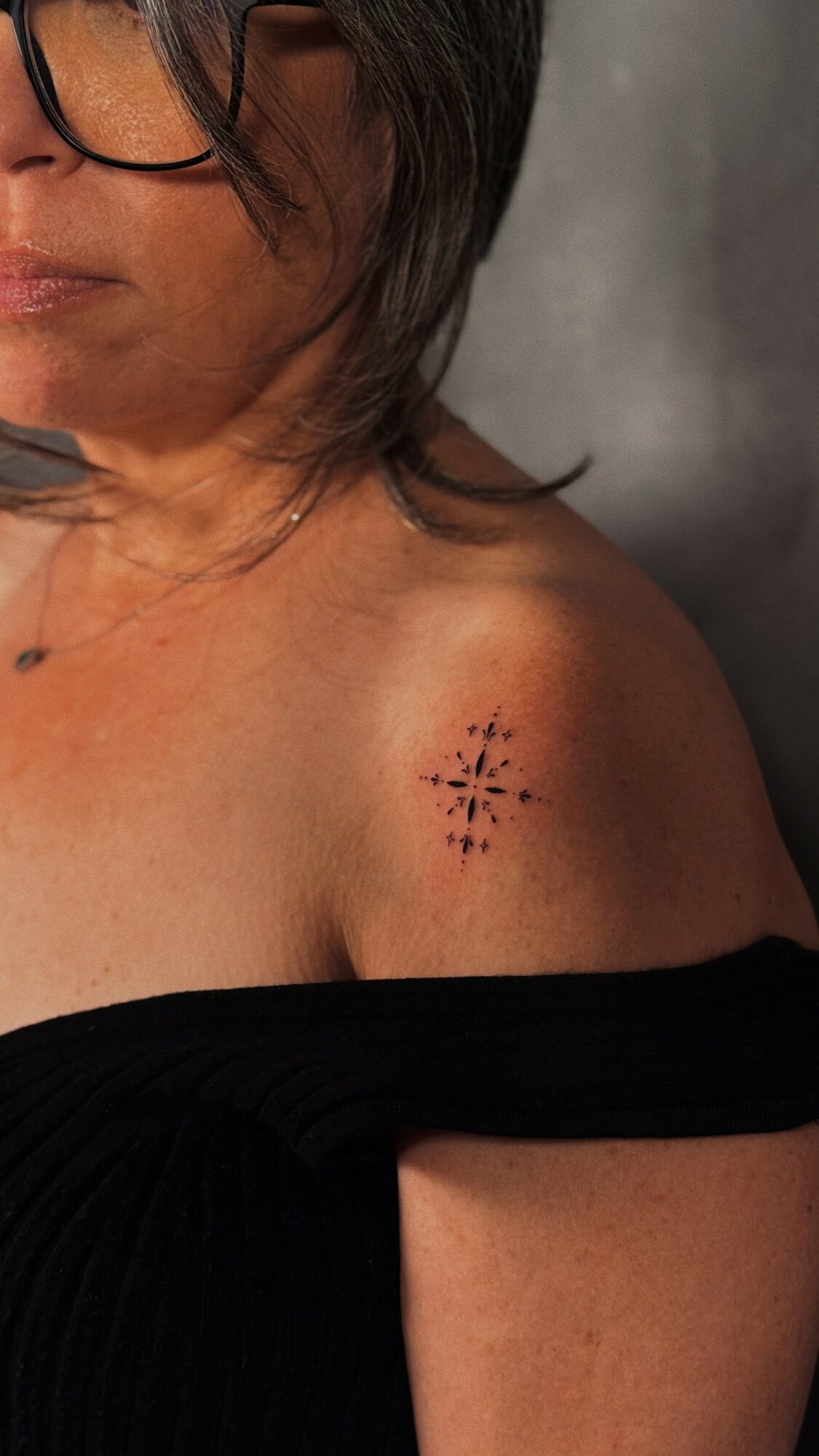 Close-up of a person's shoulder with a small tattoo of a starburst design, wearing glasses and a black off-shoulder top.