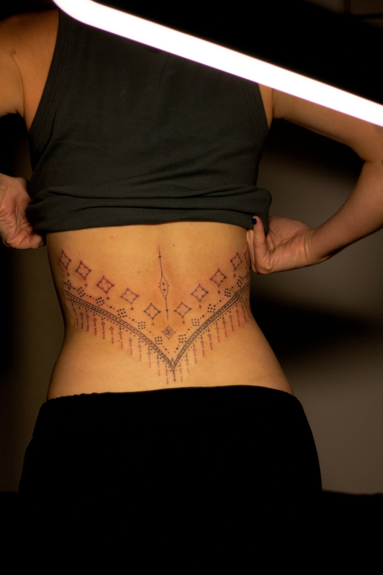 Person with a tattooed waist wearing a black top, with their hand on their hip, against a dark background.