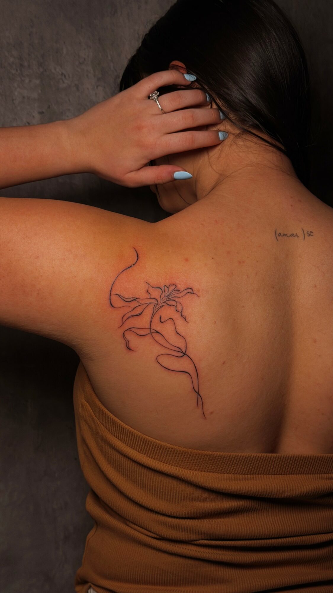 Person with dark hair, back turned, showing a colorful abstract tattoo on shoulder blade, hand touching hair, wearing a brown top.