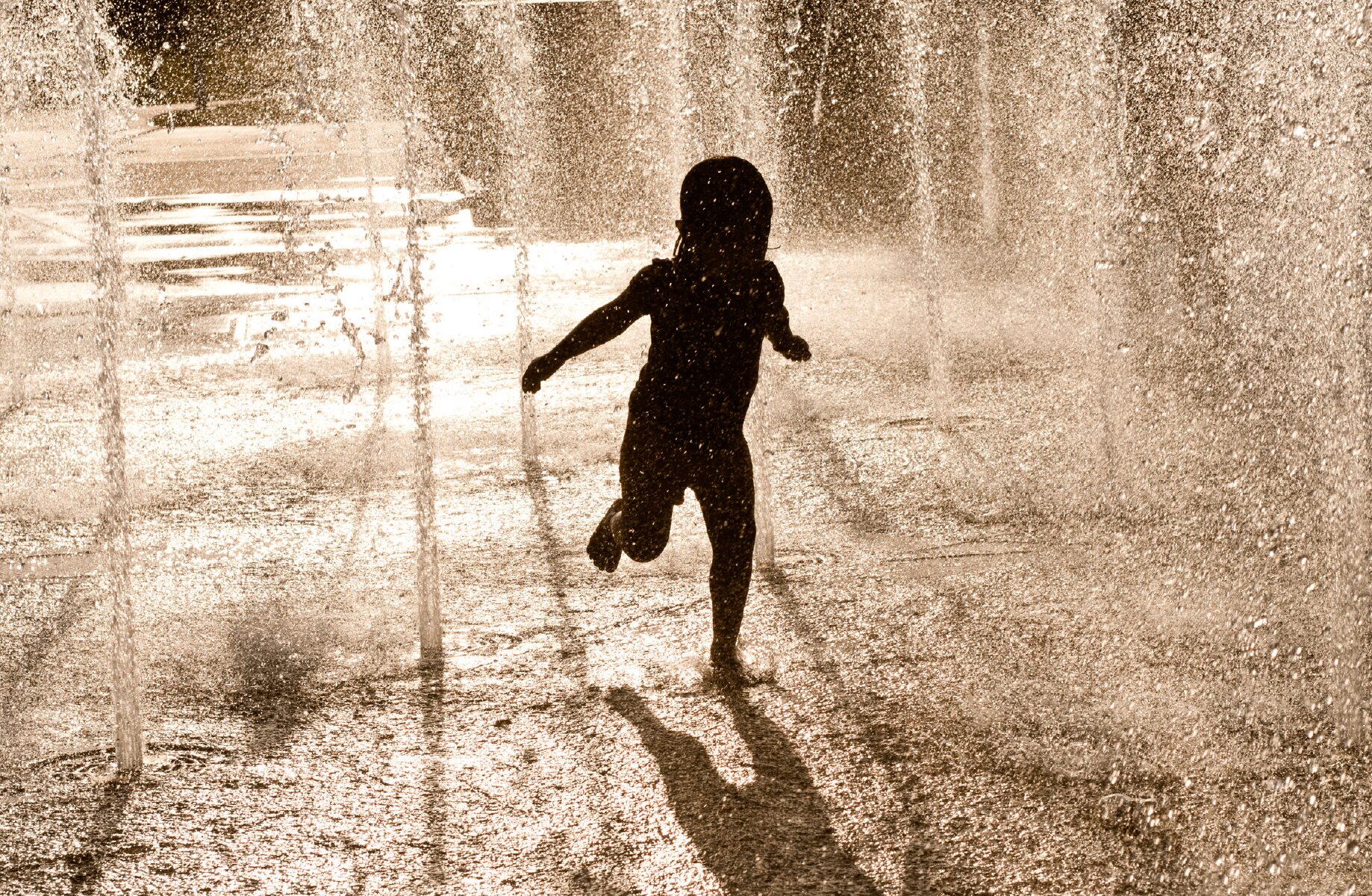 Child running through water spray with sunlight, shadow visible on wet ground, water droplets in air, outdoors.
