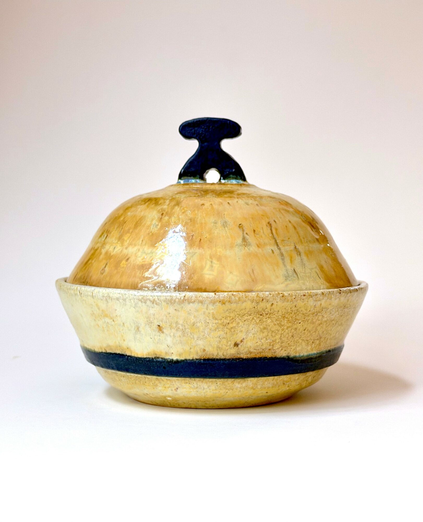 Round ceramic container with a lid and handle, beige with dark stripe, on a plain background.