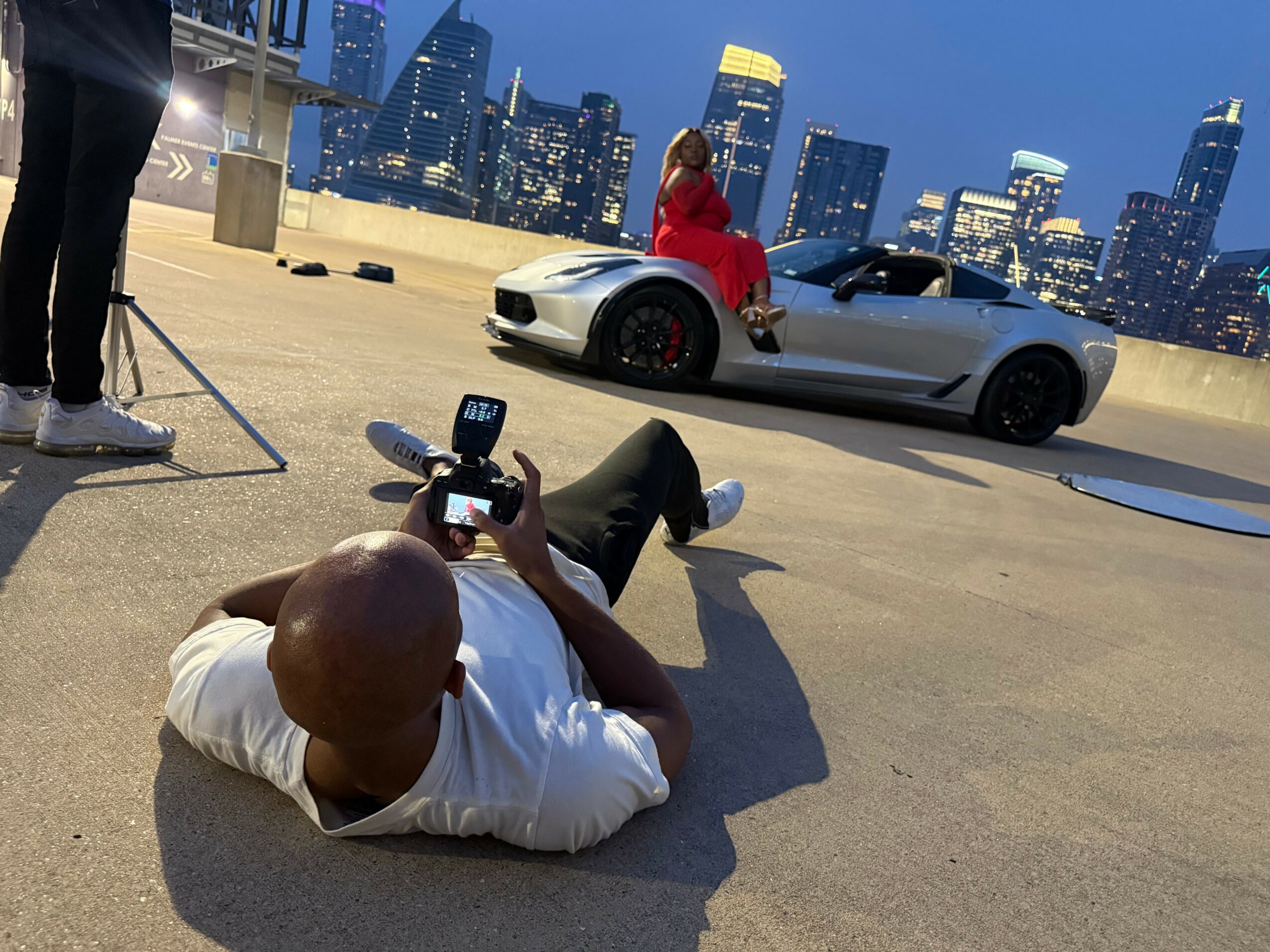 Person lying on the ground taking a photo of a woman sitting on a white sports car with a city skyline in the background.