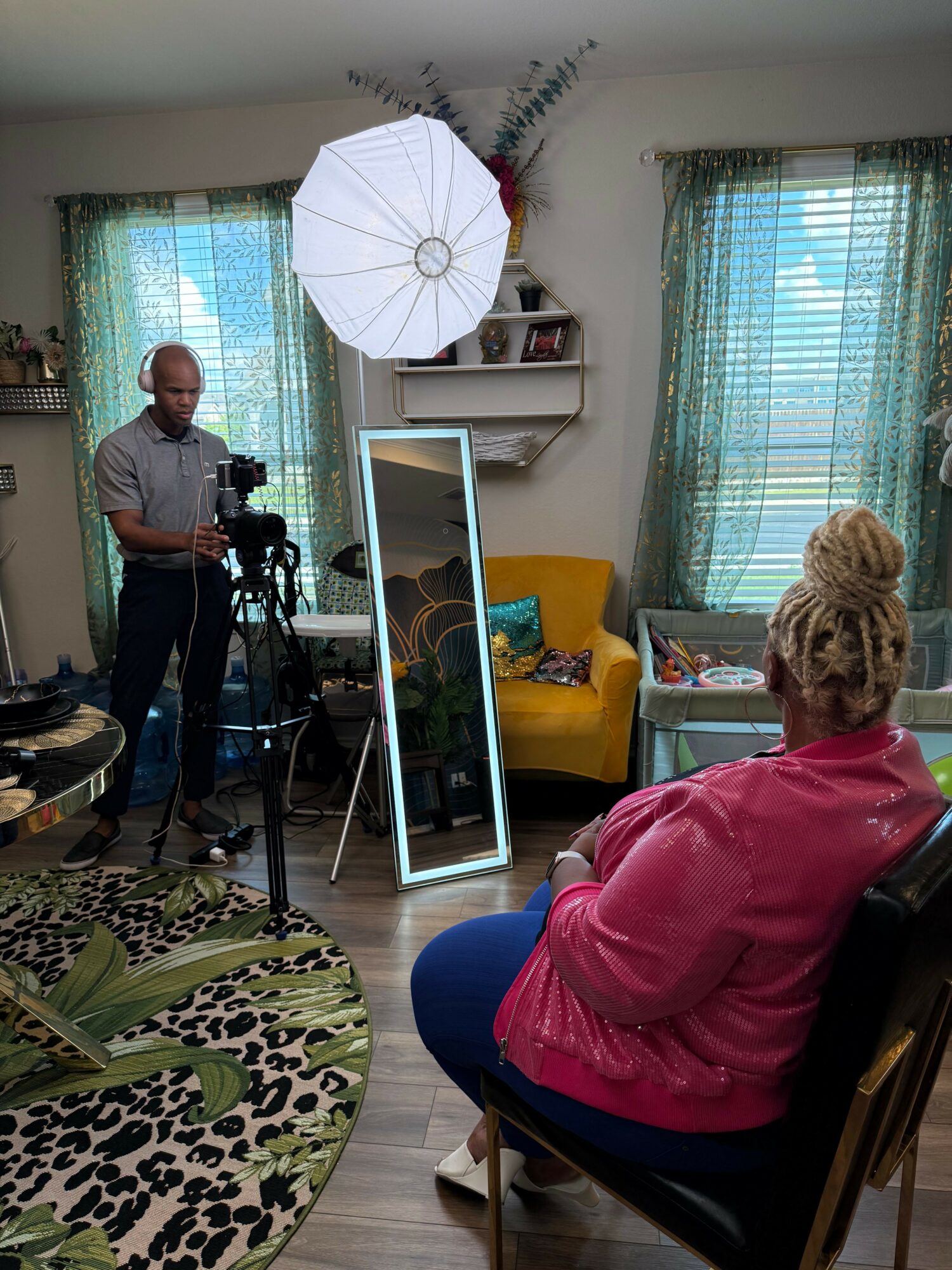 Woman in pink sitting at a table, person in gray standing near a mirror, window with blinds, and a white umbrella in a room.