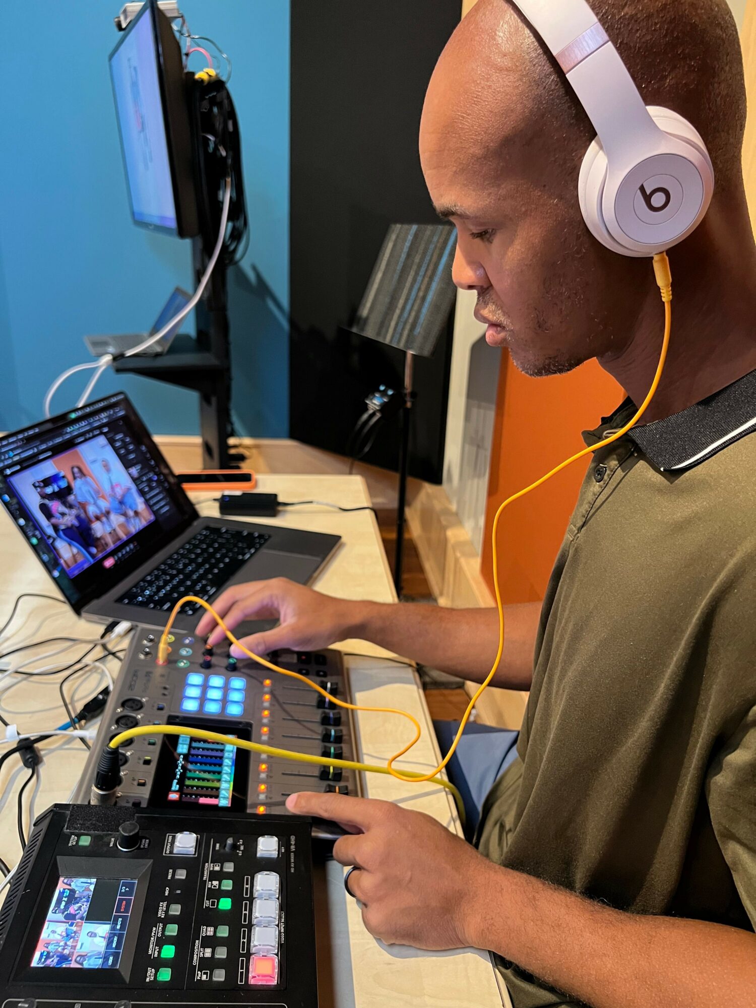 Person wearing headphones interacts with electronic music equipment on a table, with another person assisting. A tablet displays a video.