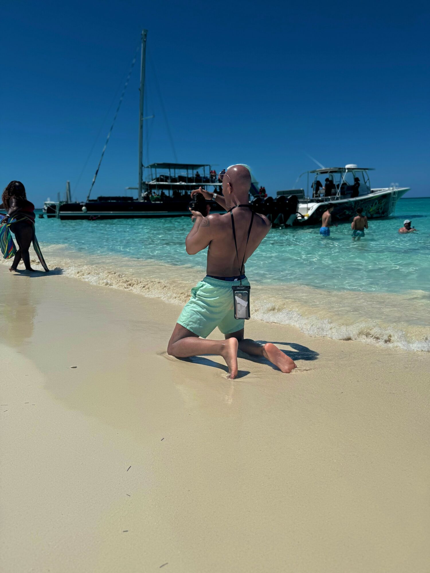 Person kneeling on sandy beach taking a photo with a camera, boats anchored in clear water, blue sky.