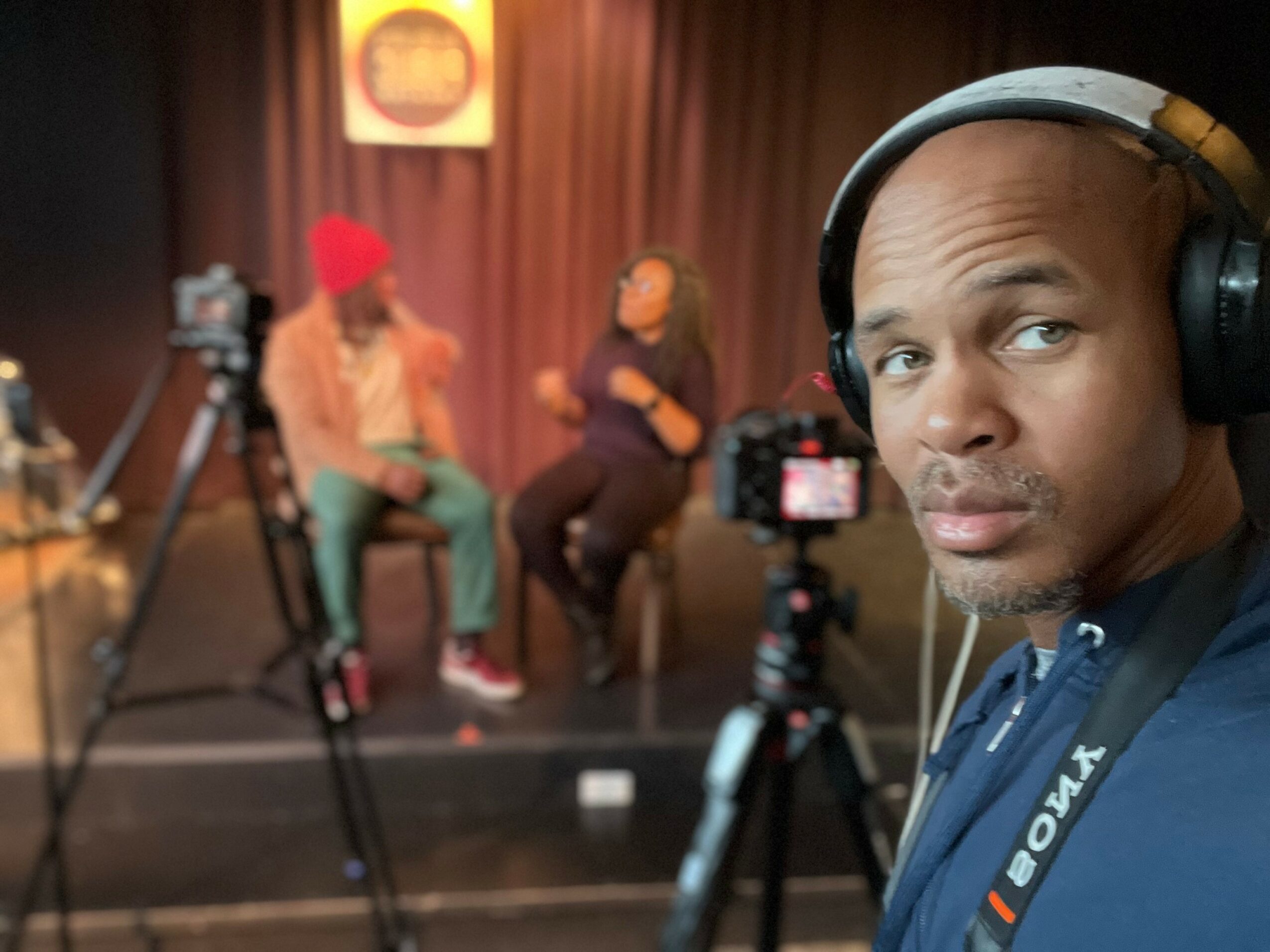 Man with headphones in foreground, two people talking on a stage in background, cameras set up, blurred background.
