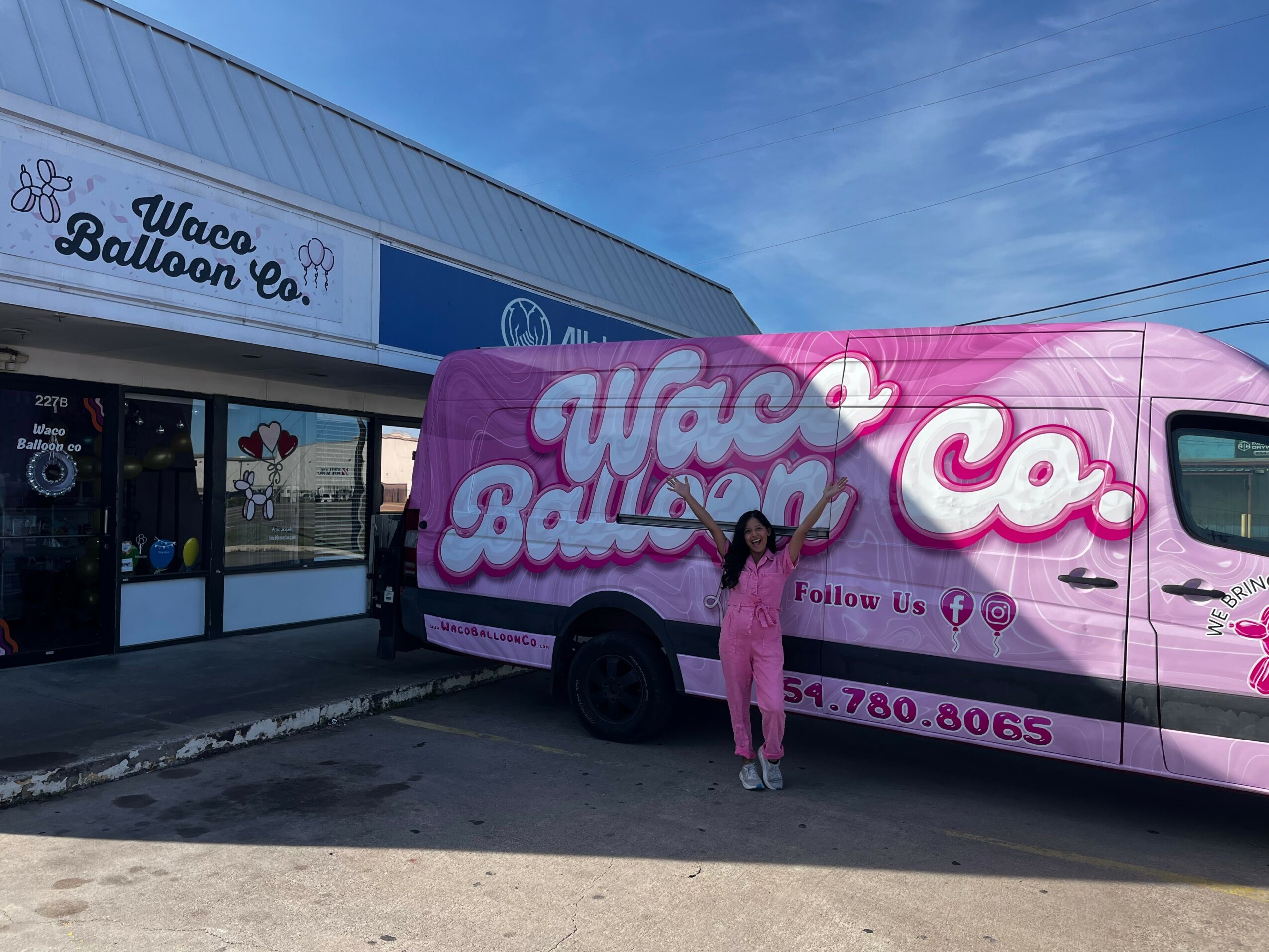 Person standing in front of a pink balloon company van and storefront, both displaying the company's logo and contact information.