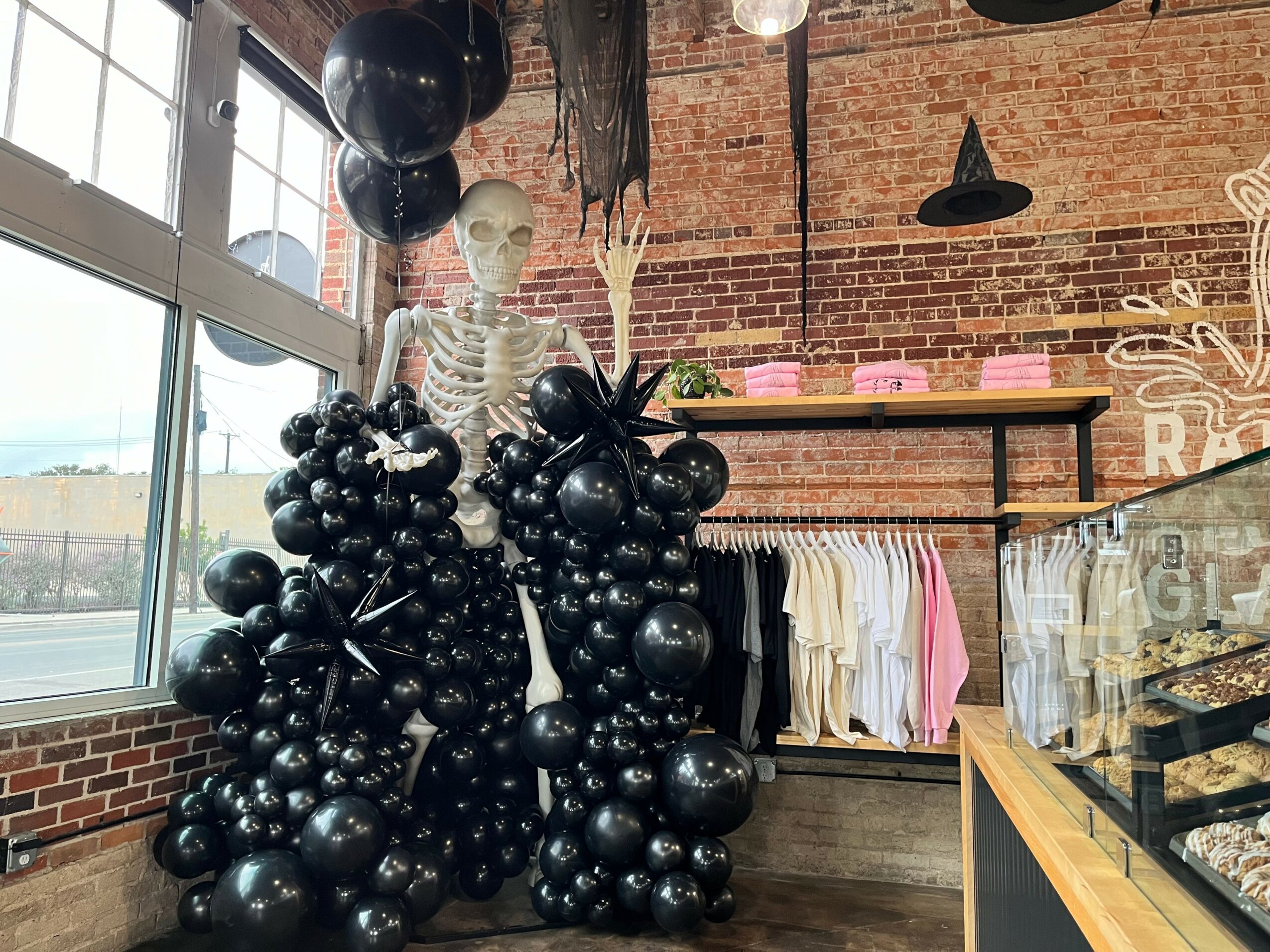Skeleton figure decorated with black balloons stands inside a room with brick walls and large windows, Halloween decorations visible.