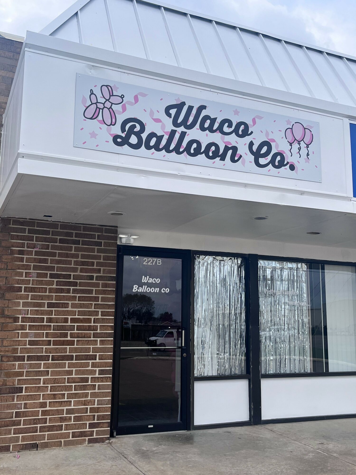 Storefront with a sign reading 'Ulcece Bawcom' and a glass door and window, brick wall at the bottom, and a sidewalk outside.