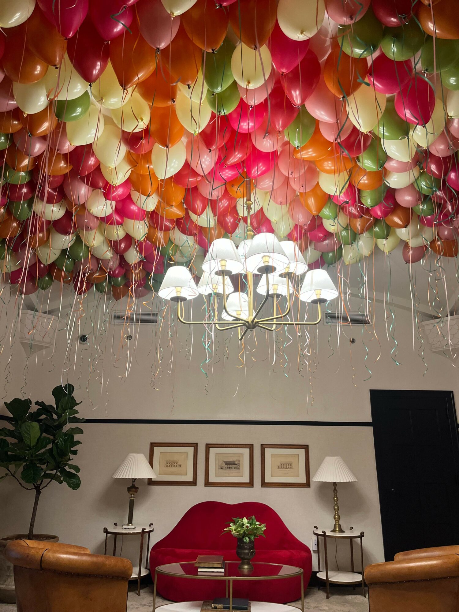 Room decorated with colorful balloons, a chandelier, framed pictures, a red tablecloth, and a potted plant.
