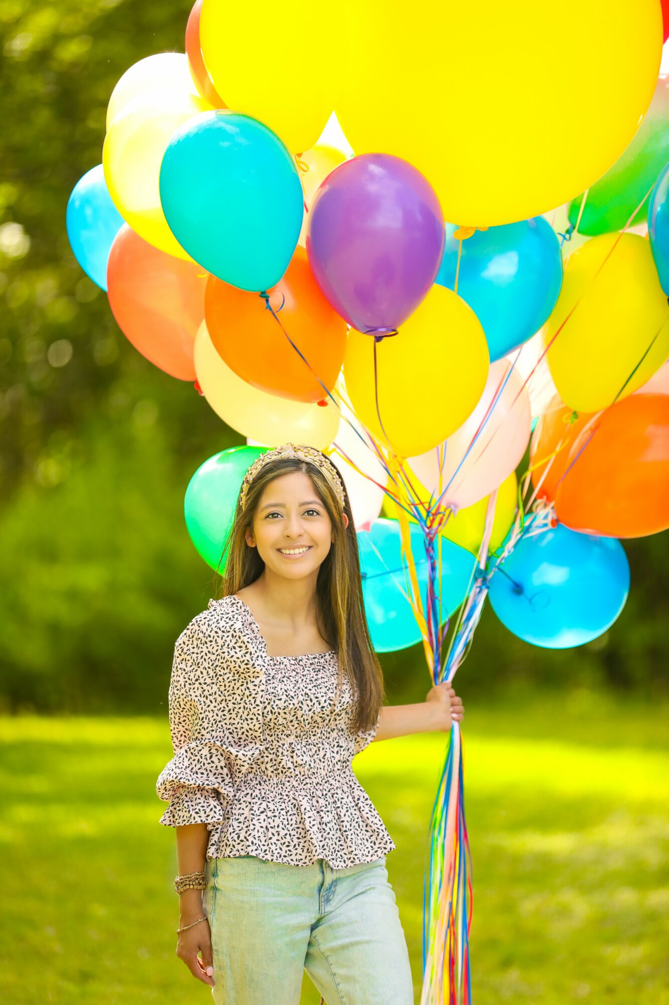 Young woman holding colorful balloons outdoors with green trees in background.