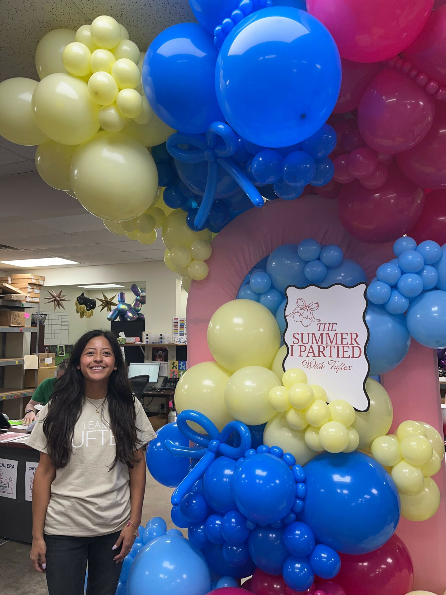 A woman stands next to colorful balloon decorations with a sign that reads 'Sprinkles & Sparkles' in the background.