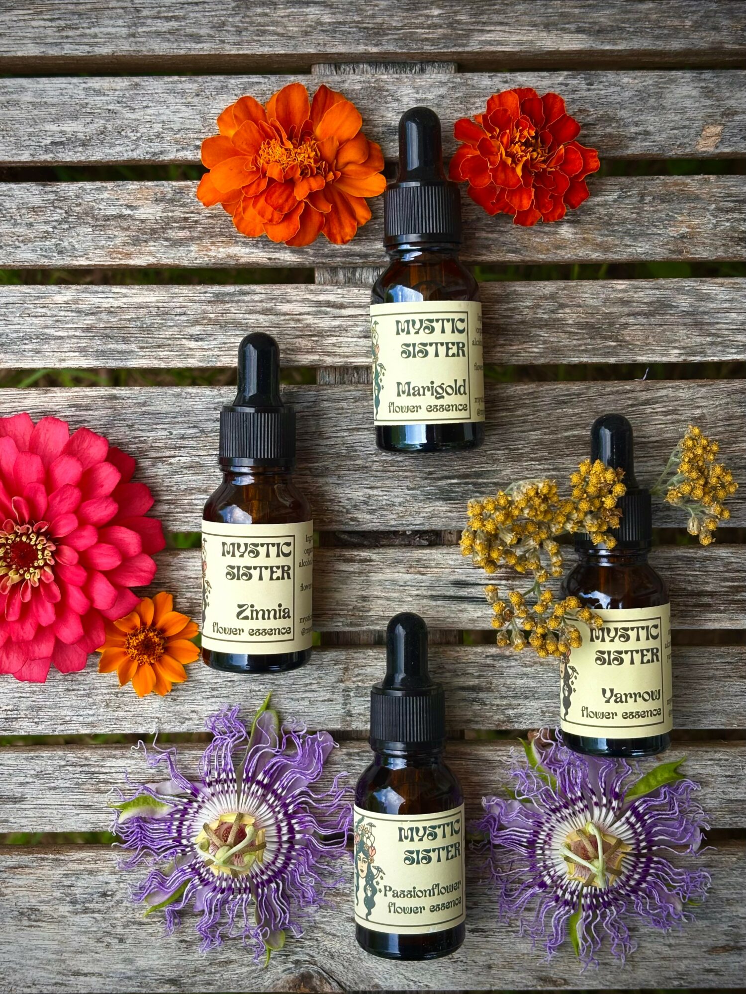 Five small bottles with dropper caps surrounded by colorful flowers on a wooden surface.