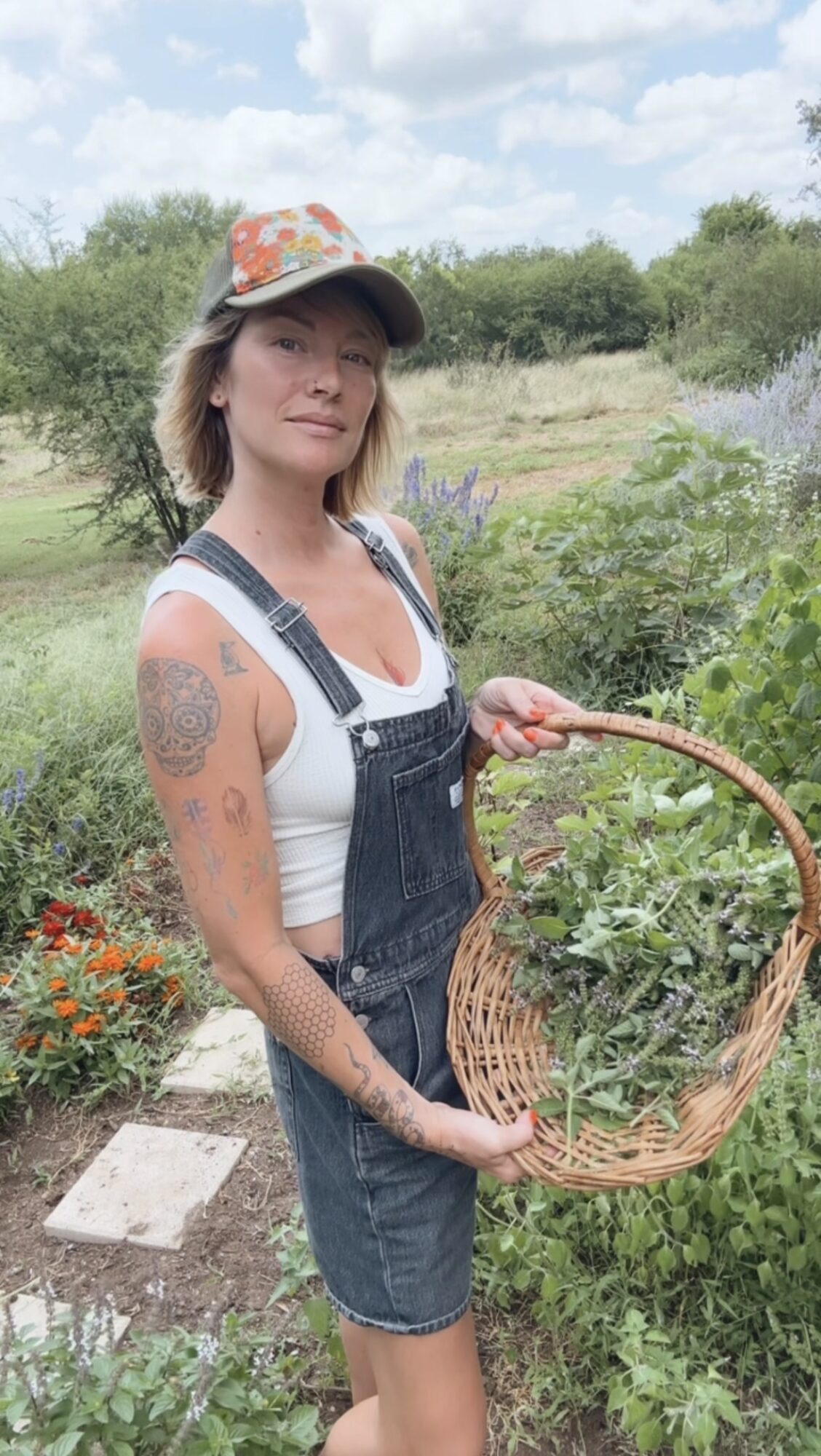 Woman holding a wicker basket outdoors with greenery and flowers, wearing a hat, white shirt, and denim overalls.