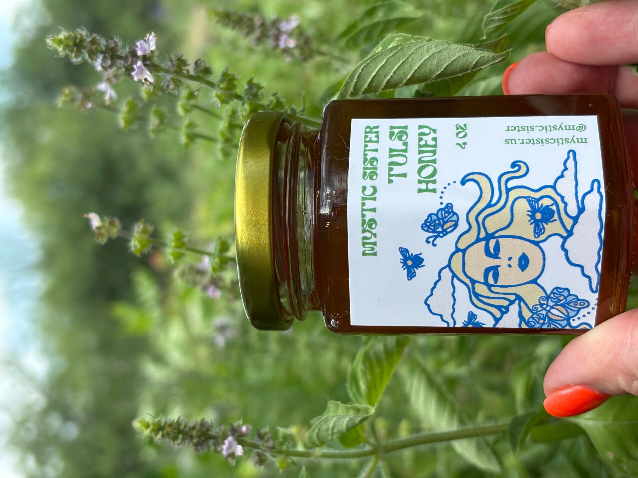 Hand holding a small jar with a label, surrounded by green leaves and purple flowers, outdoors.