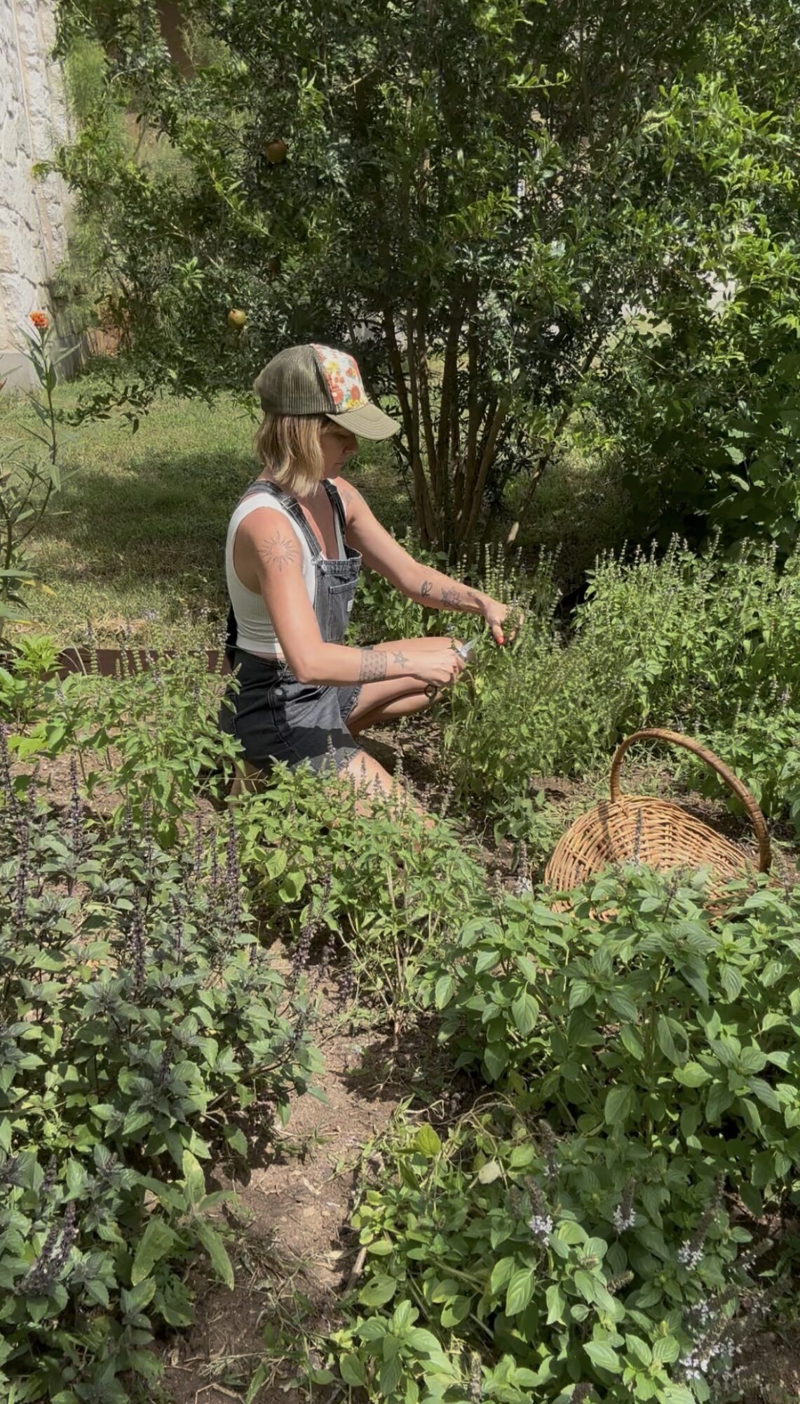 Woman in a garden with lush green plants, wearing a cap, sleeveless top, and shorts, sitting and tending plants.