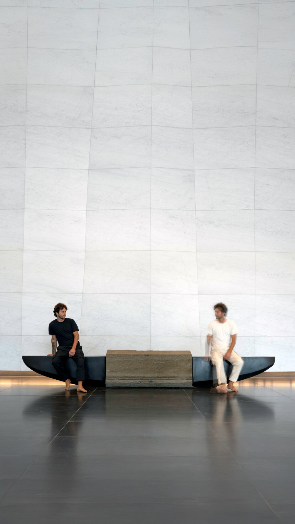 Two people sit on benches facing each other in front of a large, plain white wall.