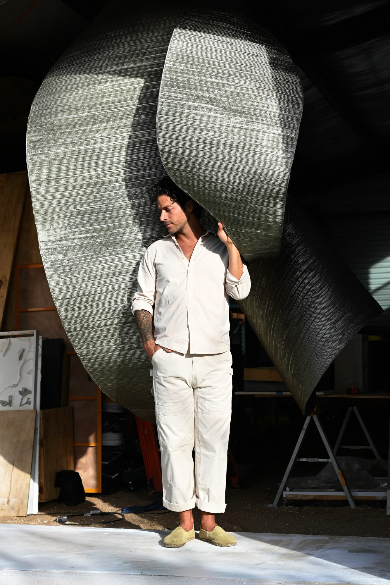 Person in white outfit standing in front of large abstract metallic sculpture, holding hair, indoors.