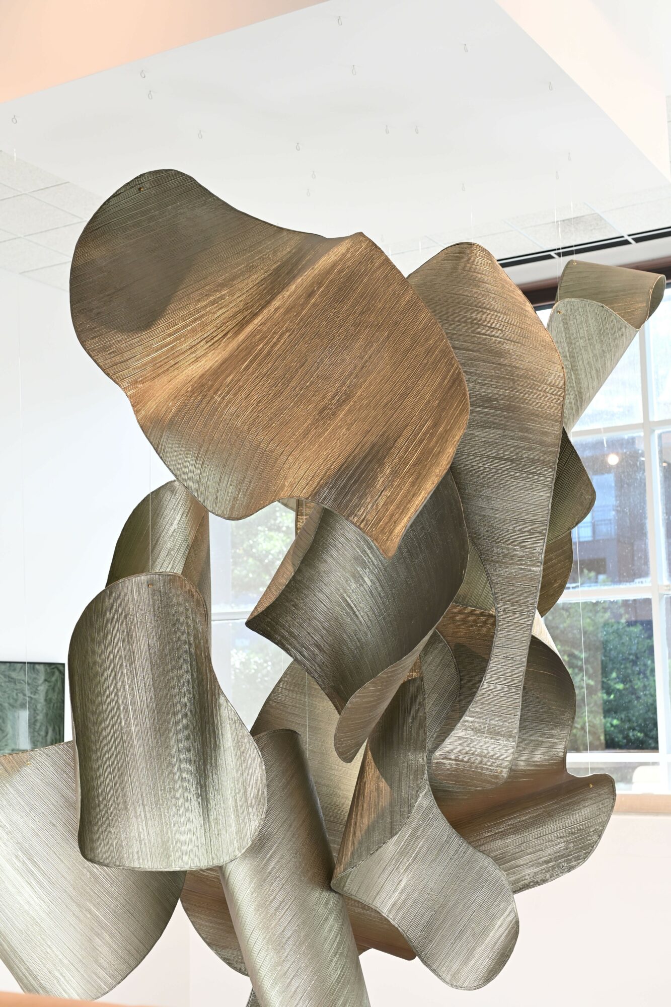 Abstract wooden sculpture with curved, overlapping shapes inside a room with large windows and natural light.