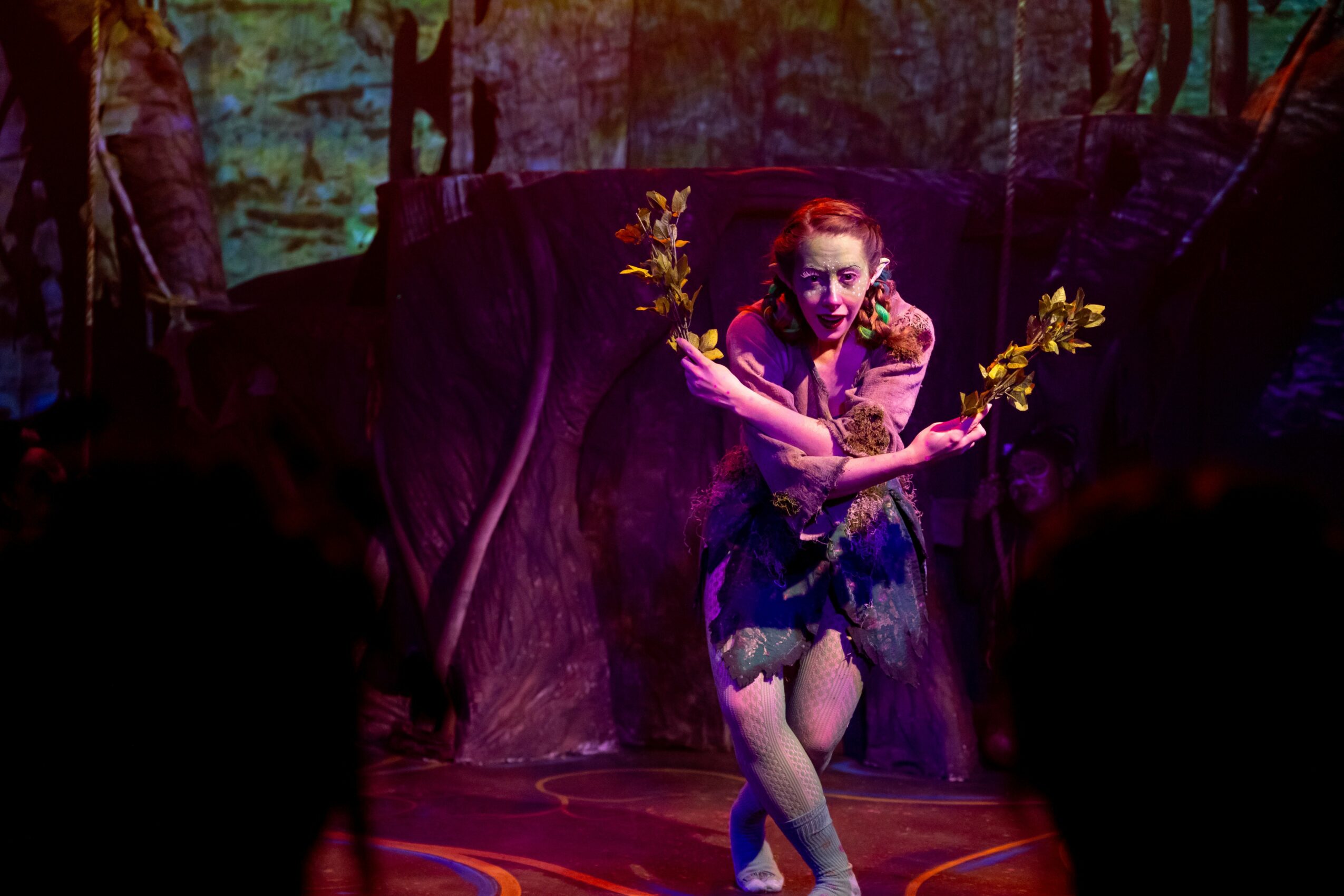 Performer with red hair and makeup, holding yellow flowers, on stage with dark background and colorful lighting.