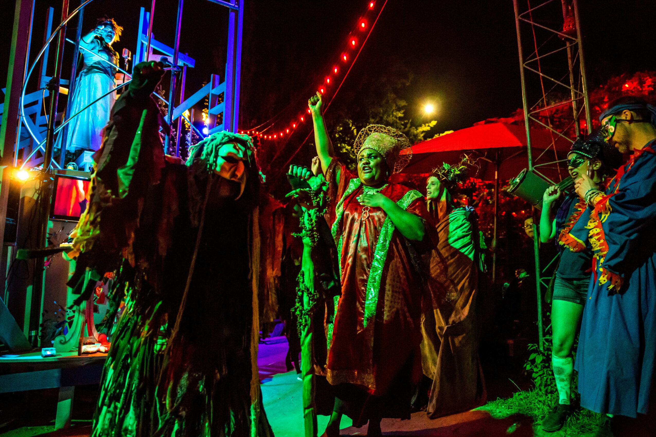 Group of performers in colorful costumes and masks on stage at night, with bright lights and a crowd nearby.