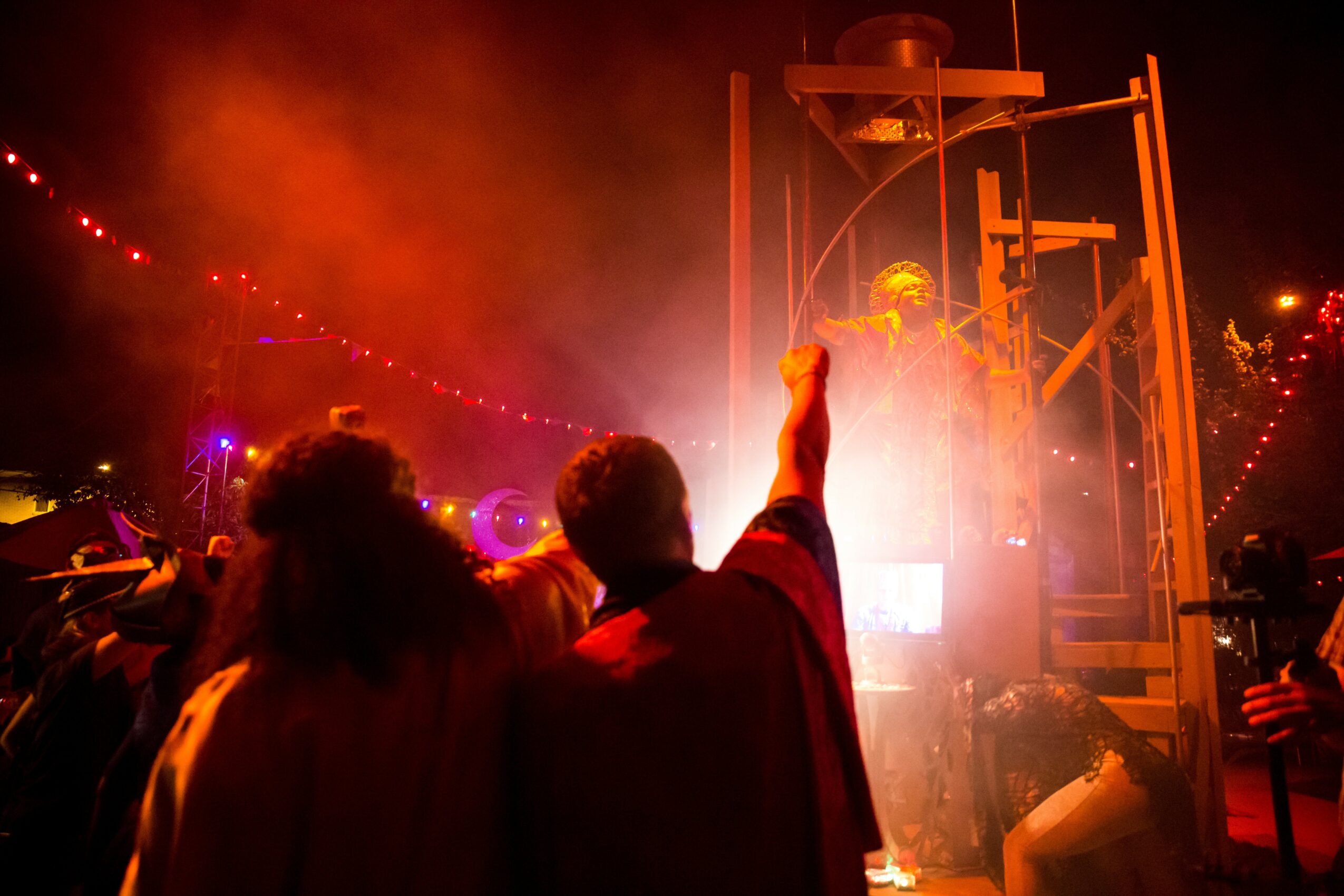 People at an outdoor event with colorful lights and a person pointing towards a structure, night setting, festive atmosphere.
