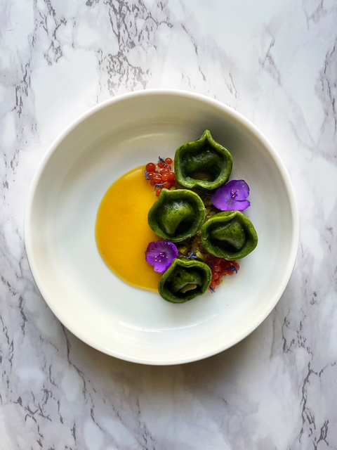 Bowl with green stuffed peppers, purple flowers, red garnish, and yellow sauce on a white marble surface.
