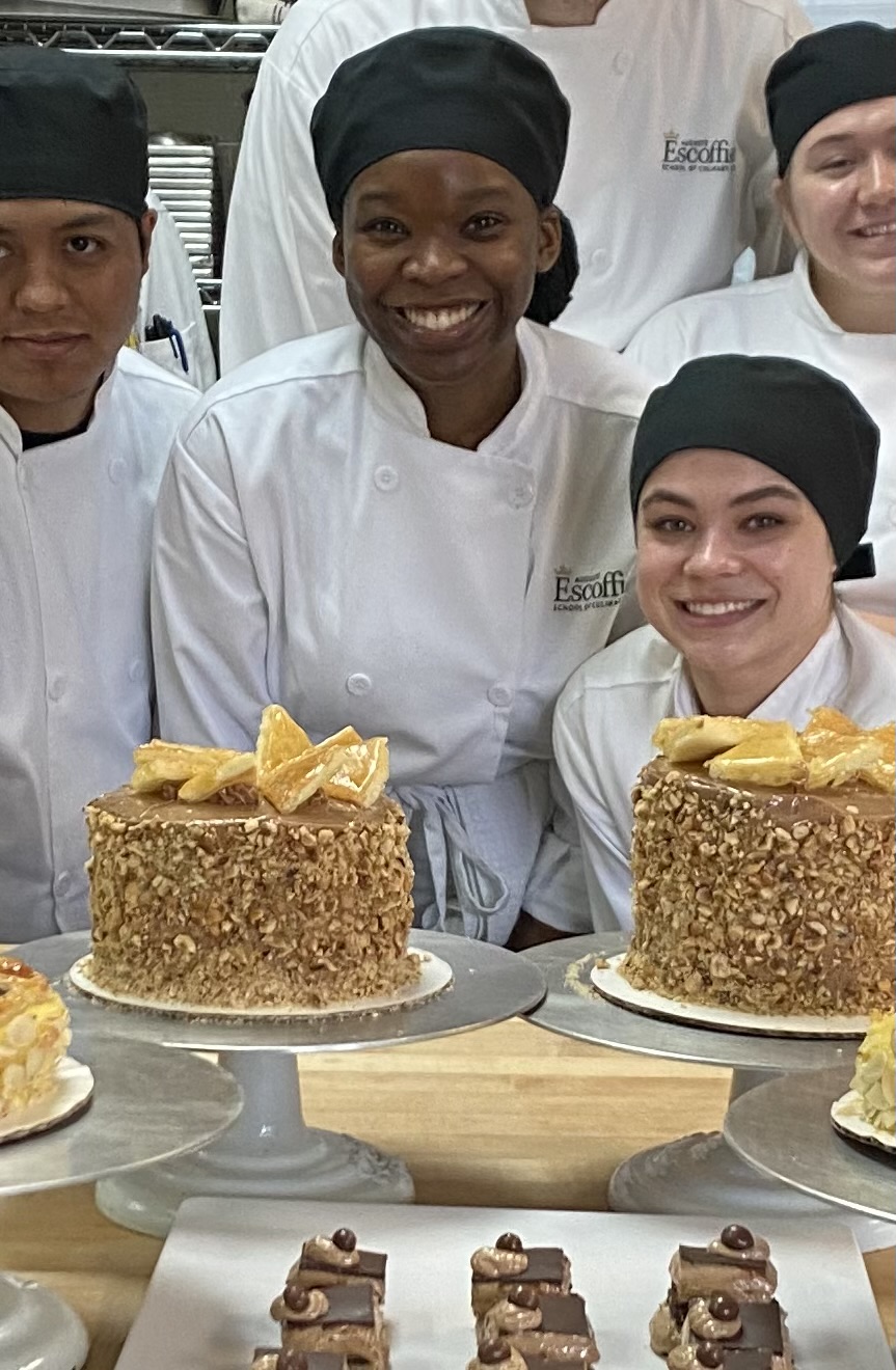 Group of smiling chefs in white uniforms and black head coverings, with decorated cakes on a kitchen counter.